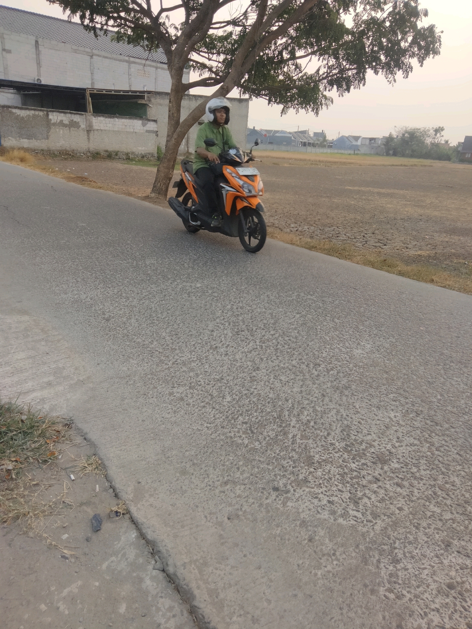 An individual wearing a green shirt and a white helmet is riding an orange scooter on a paved road. The scooter has a license plate visible. The road appears to be in a semi-urban or rural area. To the left of the road is a low wall, a building with a light-colored facade, and a tree with sparse foliage. Beyond the road, to the right, is a large, dry field with some scattered vegetation and residential buildings in the distance. The sky is hazy, suggesting a dry or dusty atmosphere. The scene appears to be in the city of Suka Sari, Indonesia.