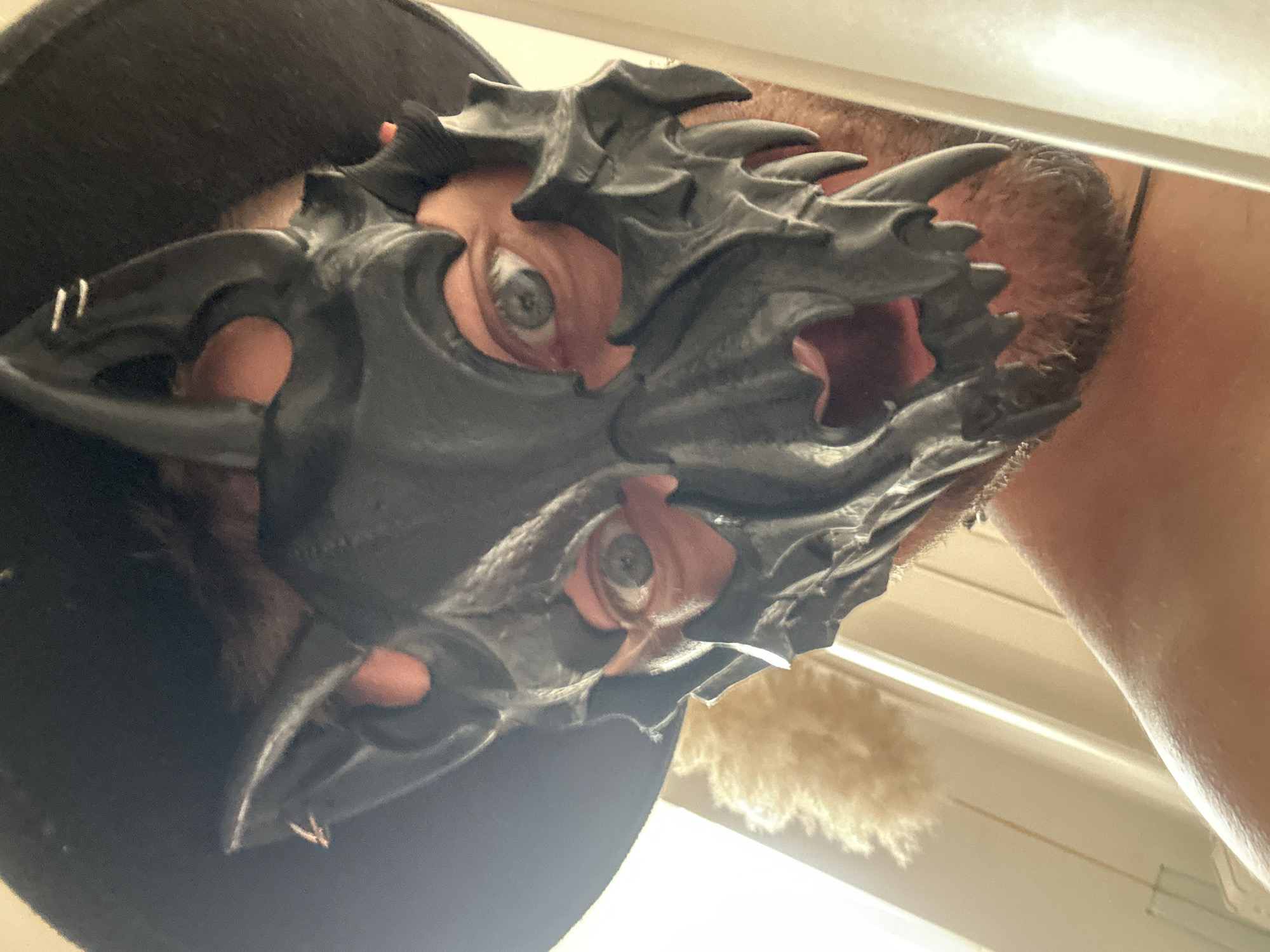 The image shows a close-up, low-angle shot of a person wearing a dark, spiky, dragon-like mask. The mask covers the upper half of the face, with visible eyes peering through the eyeholes. The person's skin tone is visible around the eyes and on the neck, with some stubble present on the chin. The person is wearing a dark t-shirt or similar garment. The background is blurred and appears to be an indoor setting with warm lighting, possibly a ceiling or wall with some light fixtures. A fluffy, light-colored object is visible in the lower right quadrant of the image, out of focus. The overall impression is that of someone posing or playing around with a costume mask, possibly indoors. The perspective creates a slightly unsettling or dramatic effect. There is no visible text in the image.