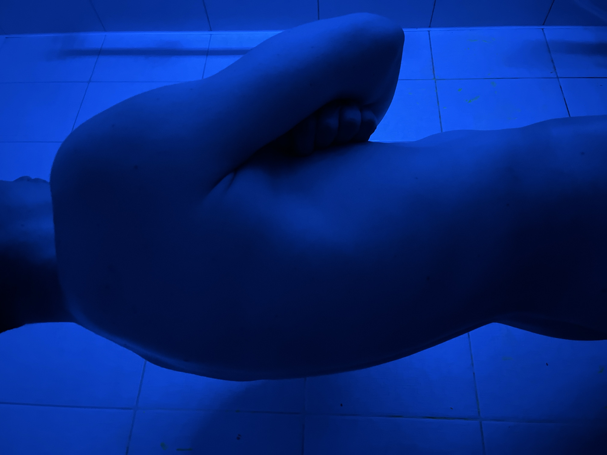 The image depicts a person lying on a tiled floor, illuminated by blue light. The person is positioned with their arm bent, the hand near the face. The tiles are square and light blue, with minimal staining. The location is not identifiable as Chaikovsky, Russia, based solely on the image.