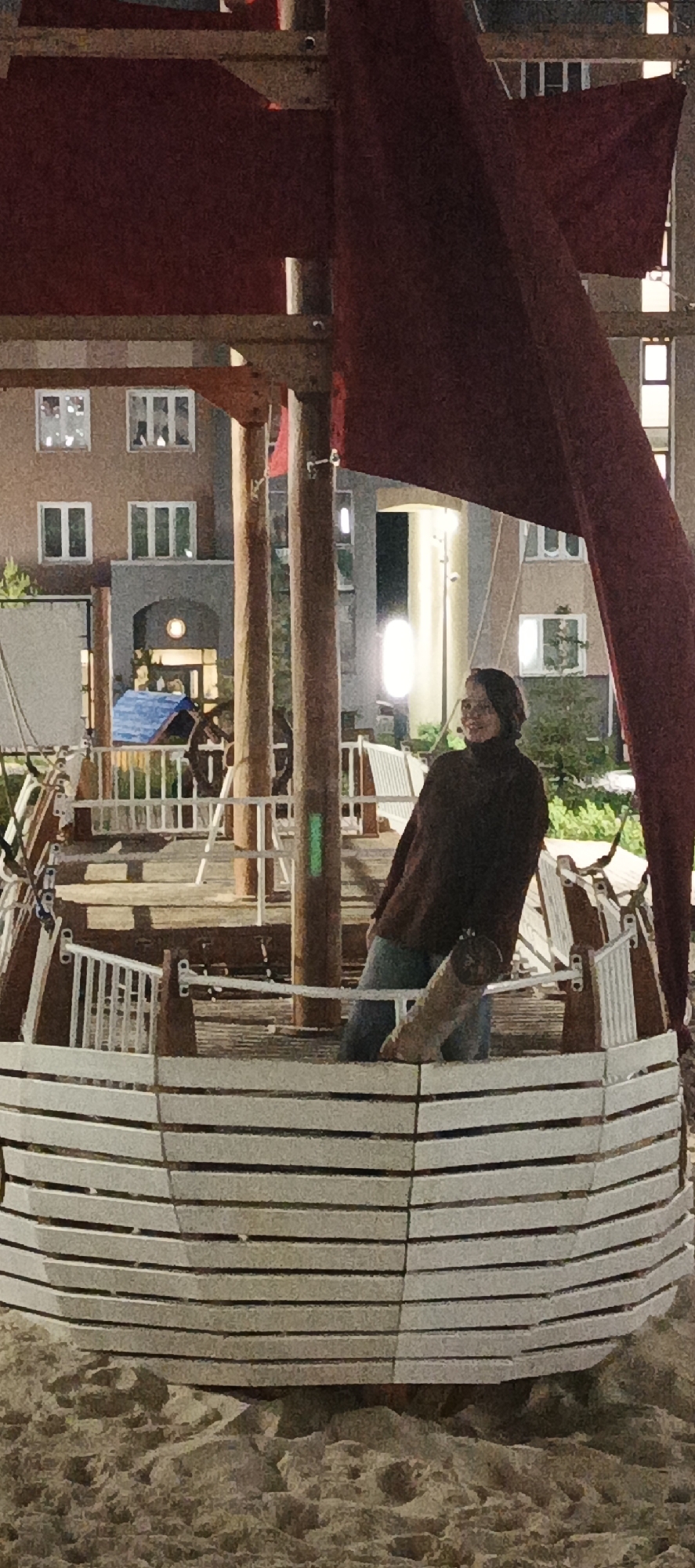 This image captures an outdoor night scene in Pervomaysky, Russia, centered on a large, boat-themed playground structure. An adult female is positioned within the wooden boat structure, standing on its deck. She wears a dark brown long-sleeved top and blue jeans, with her head turned slightly to her left, exhibiting a subtle smile. The boat structure features white, horizontally slatted hull sections, internal white railings, and multiple wooden masts supporting maroon fabric elements resembling sails or awnings. The base of the boat rests directly on a sandy or gravelly ground. In the mid-ground, behind the boat, other playground elements are visible, including a small blue-roofed structure, a large wooden ship's wheel, and additional white railings. The background features multi-story residential buildings; one on the left is light brown with several illuminated windows, and another to the right is lighter grey, also with illuminated windows. The scene is artificially lit by various sources, including bright outdoor lamps positioned between the play area and the buildings, and light emanating from the building windows. Sparse green vegetation is visible around the base of the buildings and behind the play equipment.