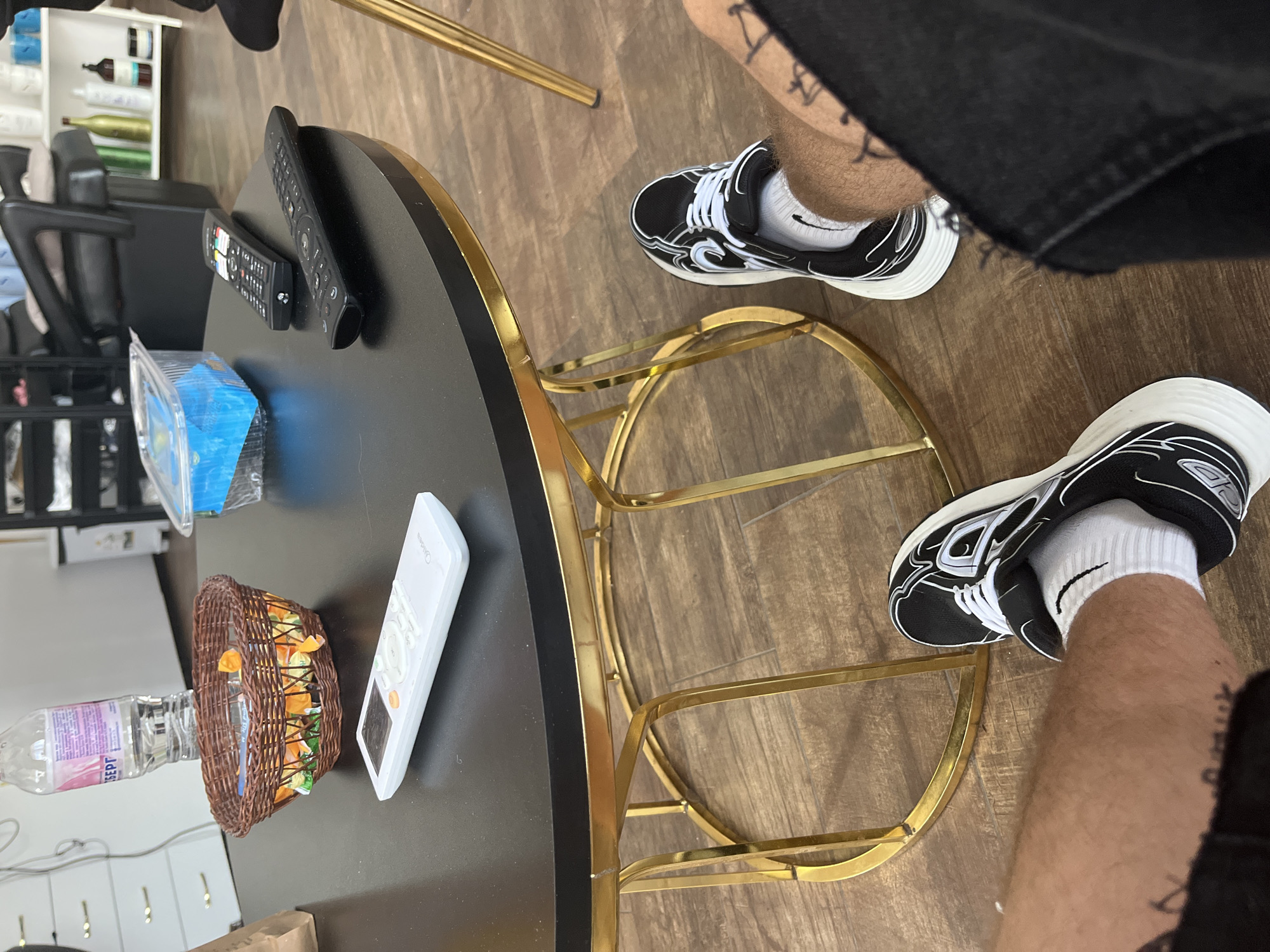 The image presents an indoor scene located in Zlatograd, Bulgaria. A low, round coffee table with a black surface and a gold-colored metal frame occupies the central foreground. On the table, from left to right, are a partially filled clear plastic water bottle with a visible "БЕРГ" (BERG) label, a small woven brown basket containing multiple candies in orange wrappers, and three remote controls. One remote is white with "Midea" branding, positioned closest to the viewer. Two black remotes are located further back on the table, along with a clear plastic container holding blue contents.
Below the table, the lower legs and feet of a person are visible. The individual wears black and white athletic shoes with white laces and white ankle socks. Tattoos are present on both legs, which are covered in body hair. Dark fabric, consistent with shorts or trousers, is visible on the upper right leg.
The background shows a light brown wooden floor. To the left, white cabinets or drawers are partially visible. In the mid-ground, a black professional salon chair with a headrest and armrests indicates a salon or barbershop setting. To the right, shelves display various bottles and containers, characteristic of personal care products found in such establishments. No direct actions or interactions are depicted beyond the person's seated posture.