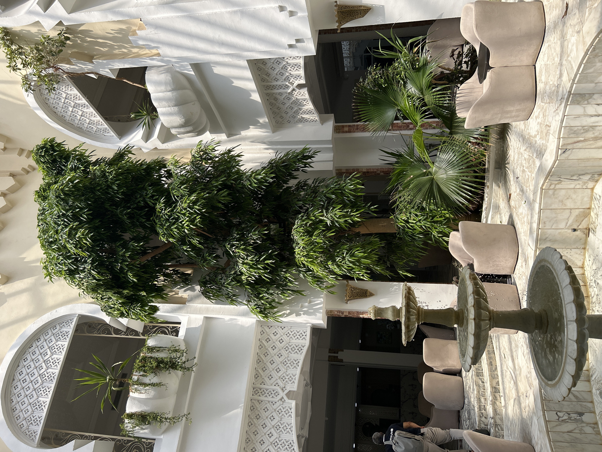 The image depicts an interior courtyard of a building in Hiboun, Tunisia. A large, leafy tree is the central element, reaching towards the top of the frame. The architecture features white arched windows with intricate lattice work, and potted plants are placed on ledges. In the foreground, a multi-tiered stone fountain is active, with water cascading down. Several plush, light-colored armchairs and a small dark table are arranged around the courtyard. A person wearing a backpack is visible on the left side of the image, walking away from the camera. The courtyard is paved with marble tiles.