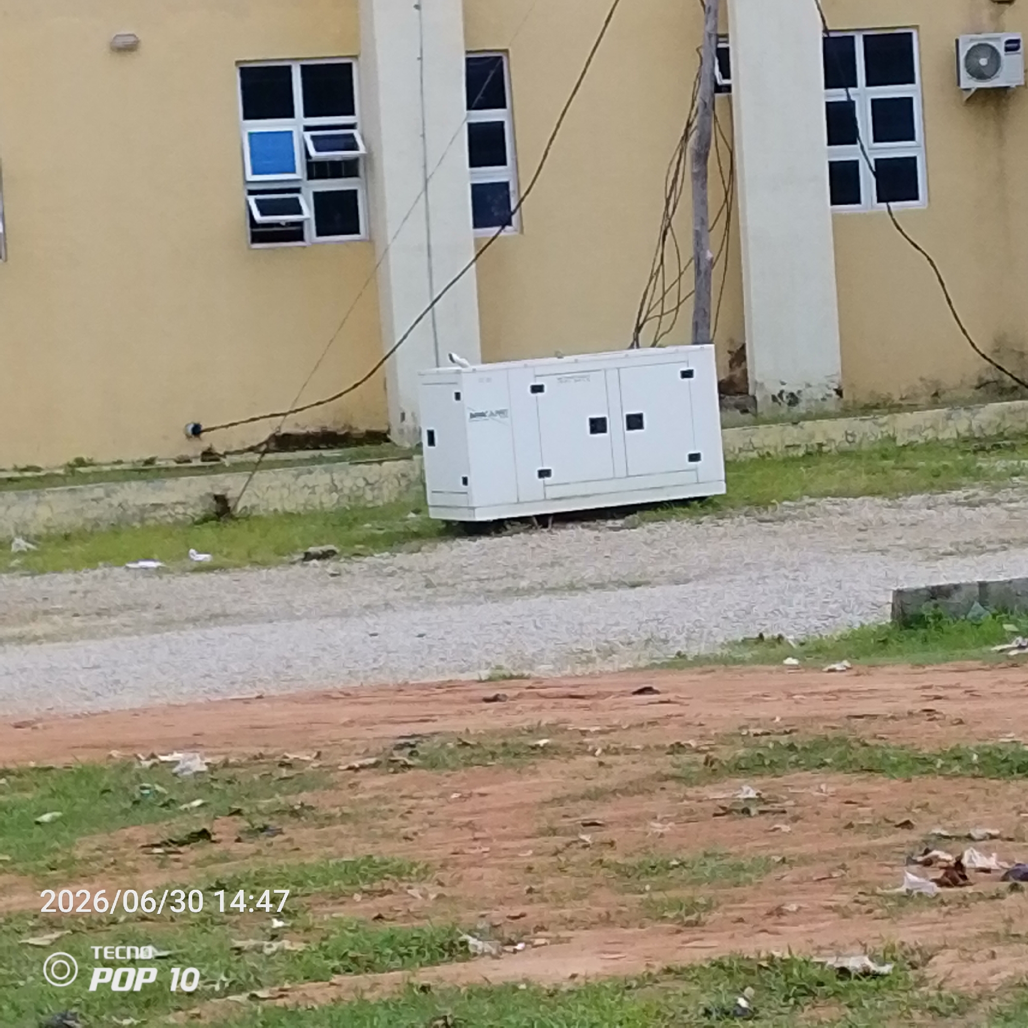 The image depicts an outdoor scene likely located in Jalingo, Nigeria, as indicated by the timestamp and phone model which often display location-specific information in their metadata. In the background, a building with a light yellow facade features several windows with dark panes. Some of these windows are partially open. To the right of the windows, a white air conditioning unit is mounted on the wall.
In the mid-ground, a large, white generator unit is situated on a gravel surface. The generator has black rectangular panels on its sides. Electrical wires, some appearing weathered and running from a wooden pole, are visible strung across the scene and near the generator. The ground in front of the generator is covered with gravel, and beyond that, a patch of green grass is visible with scattered debris.
The foreground shows a reddish-brown dirt area interspersed with patches of green grass, also containing various pieces of litter. A date and time stamp, "2026/06/30 14:47," and the "TECNO POP 10" branding are superimposed on the lower left portion of the image. No people are visible in the frame. The overall environment appears to be a functional area adjacent to a building, possibly a school, hospital, or office complex, given the presence of a generator and air conditioning unit.