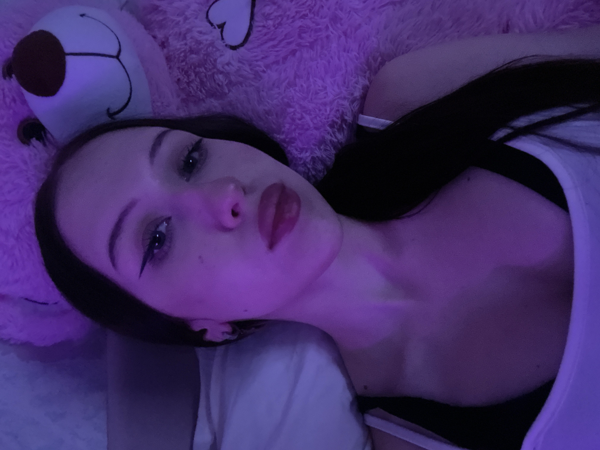 The image shows a young adult female illuminated by purple and pink ambient light, reclining in what appears to be a bedroom. Her head rests on a white pillow positioned on the left side of the frame, and her long, dark hair cascades around her shoulders. She is wearing a white tank top over a dark-colored garment. Her face is made up with prominent winged black eyeliner and glossy dark pink lipstick, and she gazes directly at the camera with a neutral expression. To her right, a large pink fluffy teddy bear is partially visible, featuring a white muzzle, a brown nose, and a stitched smile; a white heart-shaped detail is also visible on its pink fur. The immediate background consists of a plain, light-colored surface on the left and extensive pink fluffy material on the right. There are no discernible visual cues within the image to specify its location as the city of Votkinsk, Russia.