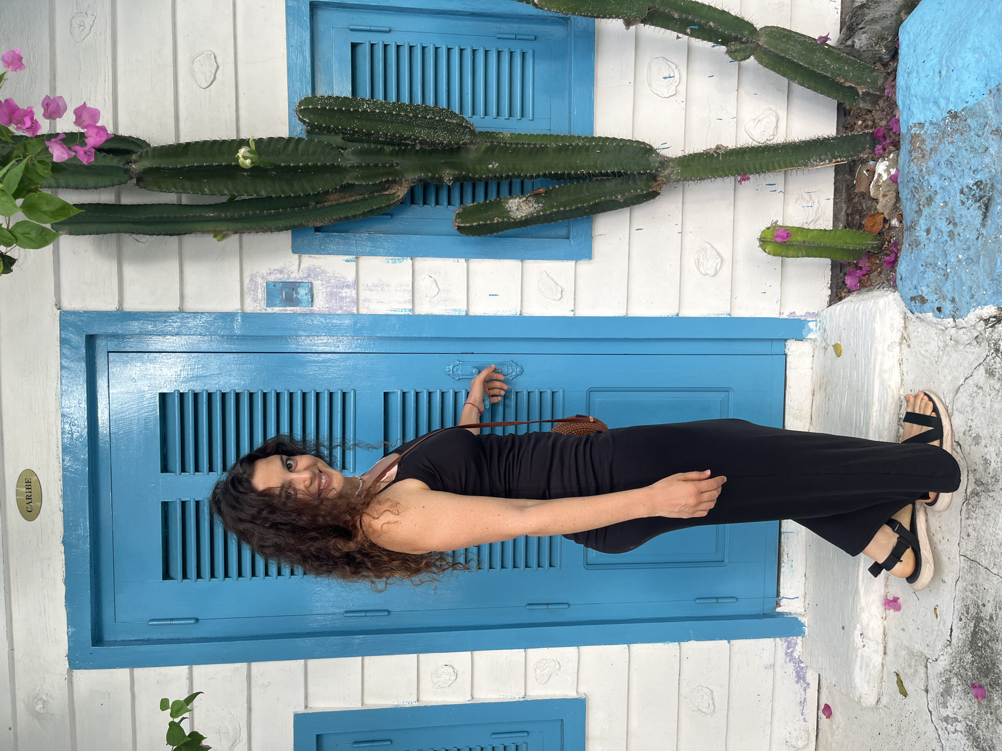 A young woman with long, curly brown hair, wearing a black tank top and black pants, stands next to a bright blue door with slatted shutters. She is looking at the camera and smiling. A small brown crossbody bag is slung over her shoulder. To the right of the door, a tall cactus with pink flowers is growing. The building is made of white horizontal wooden planks. There are also blue shutters on the right side of the image, with a small light blue box attached to the wall. The ground in front of the door is a concrete walkway with scattered pink petals. The scene is likely in a tropical or coastal location.