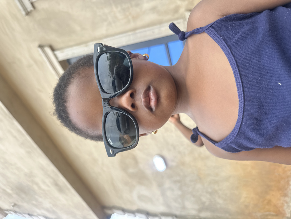 A young child is centrally positioned in the foreground against an outdoor backdrop in Ilishan, Nigeria. The child has dark skin, short dark hair, and wears black Ray-Ban Wayfarer-style sunglasses, with the brand logo identifiable on the left lens. Gold-colored hoop earrings are visible in both ears. The child wears a dark blue ribbed tank top with shoulder straps tied into knots and maintains a neutral gaze toward the viewer. Reflections in the sunglass lenses show a light-colored vehicle and ambient outdoor elements. The background features a light brown stucco or rendered building wall with decorative molding on the upper right and a blue-framed window or door. An oval-shaped light fixture is mounted on the wall to the left. A partial figure, indicated by a dark shoulder and a tied strap, is visible behind the primary subject's right side. The scene is under bright daylight conditions.