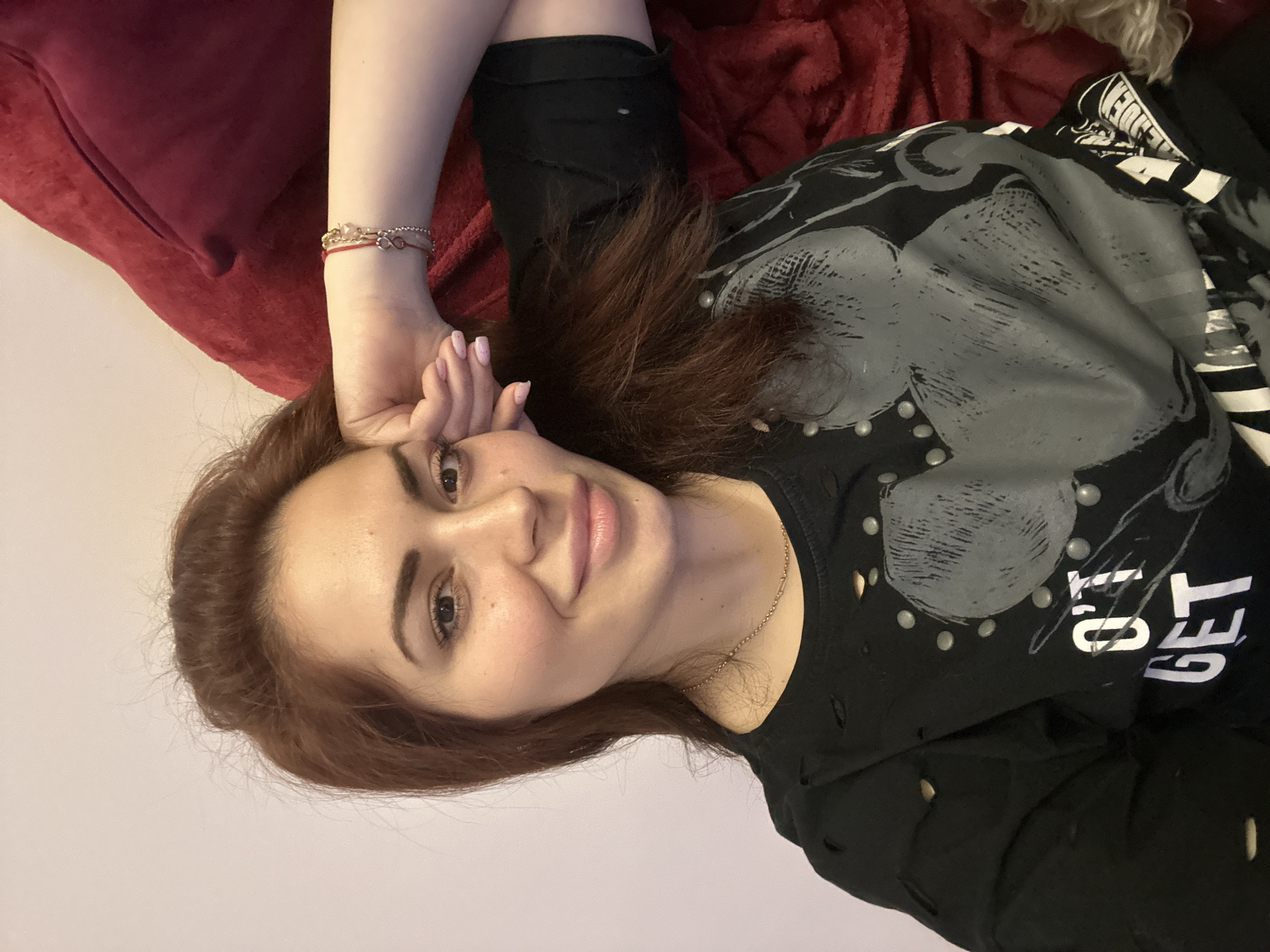 A woman lies on a bed, positioned on her side with her head tilted towards the camera. She has fair skin, brown eyes, and auburn hair. She is wearing a black t-shirt with studded accents. Her left arm is raised, with her hand near her face. On her wrist are two bracelets. The background consists of a red pillow, a light-colored wall, and a portion of the bed. The location is not discernable.