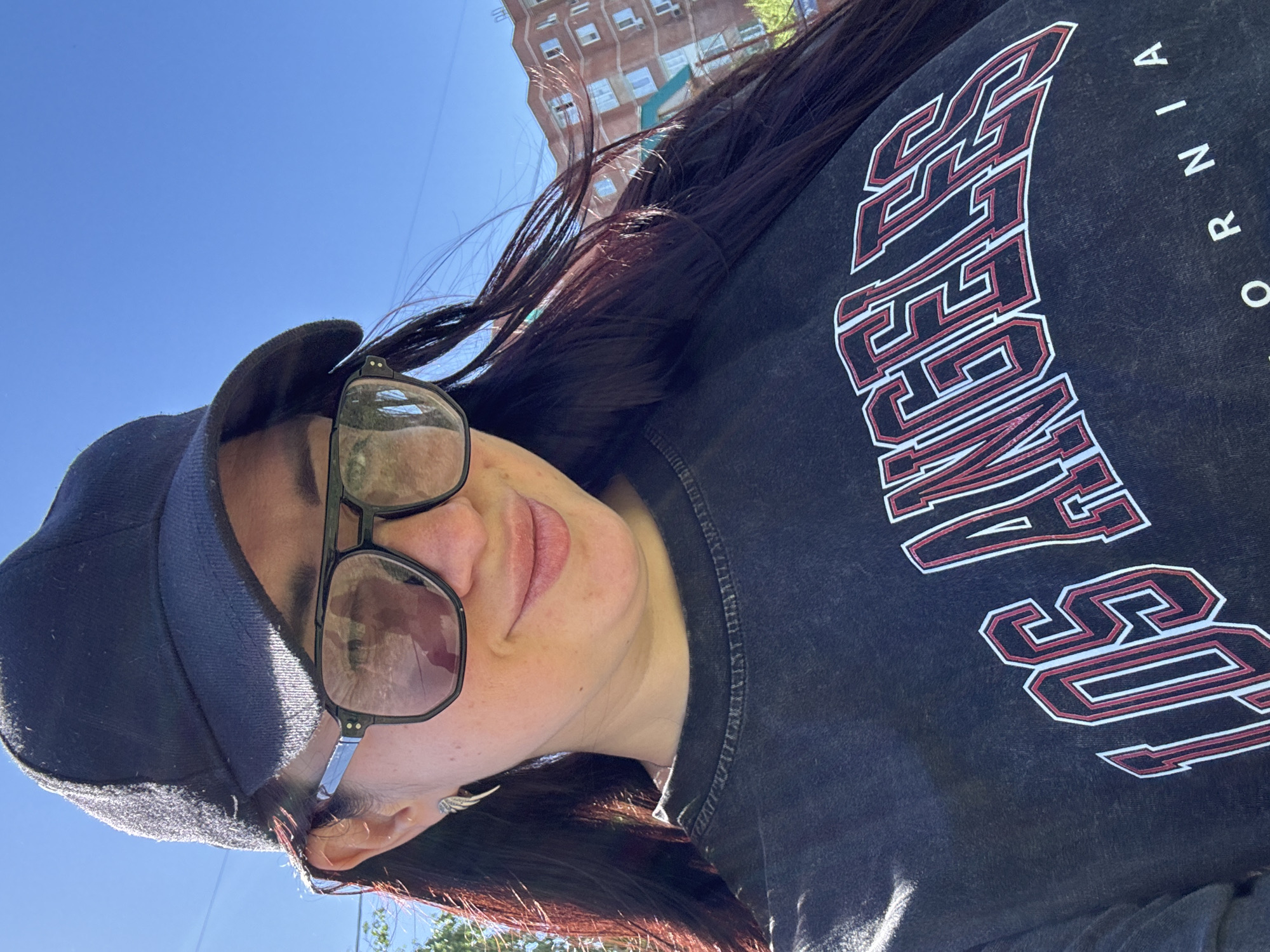 A woman wearing a dark-colored cap and oversized glasses is pictured from a low angle. She is also wearing a dark-colored shirt with the words "Los Angeles, California" in white and red lettering. In the background, there is a building and a blue sky. The location is Unknown, Unknown.