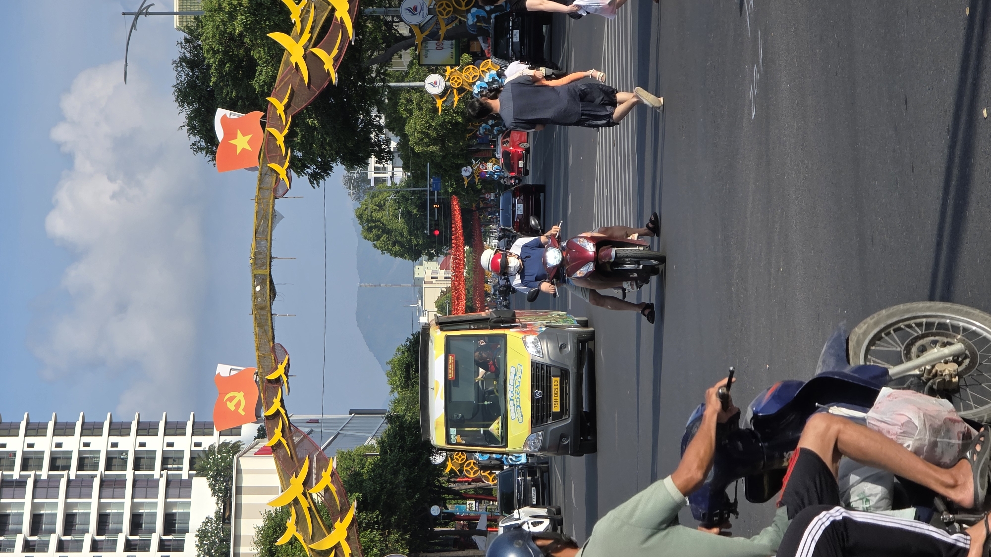 The image depicts a street scene in Nha Trang, Vietnam, with traffic, pedestrians, and decorations. In the foreground, a motorcyclist in a green shirt is visible to the right of the frame. He is stopped with his right leg on the ground and his left leg on the bike. In the middle ground, a yellow commercial vehicle with colorful graphics drives towards the viewer. A red motorbike with two riders is positioned slightly behind and to the right of the yellow vehicle. A person is walking across a crosswalk to the right of the vehicles. Overhead, a decorative structure adorned with Vietnamese flags and yellow bird-shaped ornaments spans the street. Buildings are visible on the left side of the frame and mountains in the distance. The sky is blue with scattered clouds.