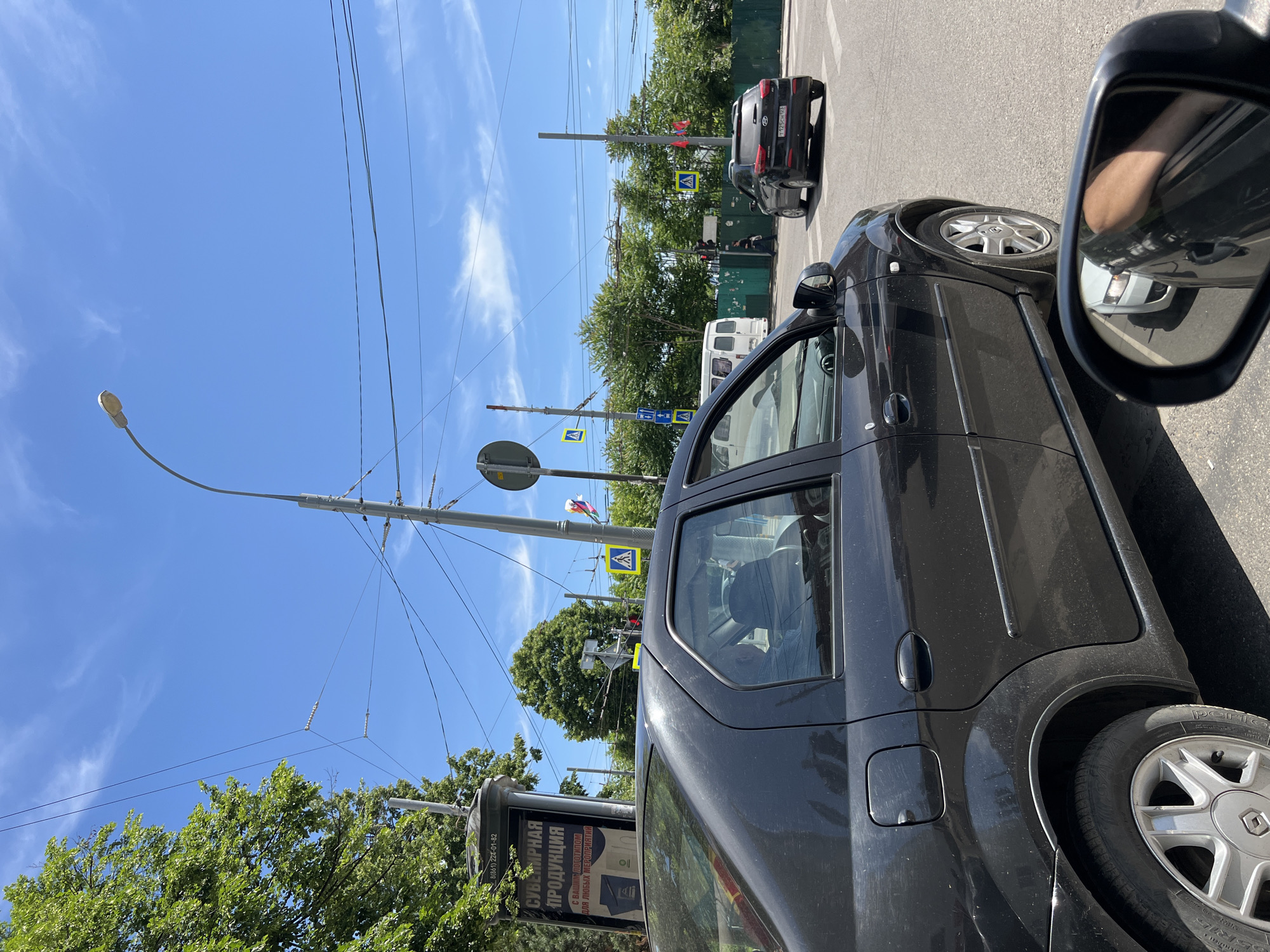 A street-level photo captures a sunny daytime scene from inside a black vehicle, likely stationary in traffic. The vehicle's right side, rear wheel with a silver Renault hubcap, and prominent side mirror are visible in the foreground. The mirror reflects a portion of the photographer's arm or hand and part of a white vehicle.
Ahead, a black sedan with Russian license plate "T125CH 93" is visible. Further down the street, a white van or minibus can be seen. The road appears to be asphalt, and to the right, a green fence borders a line of lush green trees, suggesting a park or green area.
Above, a clear blue sky with sparse white clouds dominates the upper half of the image. Numerous power lines and trolleybus wires crisscross the sky, supported by utility poles. A street lamp extends from one pole on the left.
Several road signs are present: two yellow diamond-shaped signs indicating a pedestrian crossing, and a blue rectangular sign. On the far left, a billboard or advertising stand features Russian text. "СУВЕНИРНАЯ ПРОДУКЦИЯ" (Souvenir Products) is clearly visible, along with the phrase "с вашим логотипом для любых мероприятий" (with your logo for any events) and a phone number: "(861) 224-01-02". The tire of the foreground car has "PIRELLI" written on it.
The overall scene depicts a typical city street in Krasnodar, Russia, on a bright, clear day.