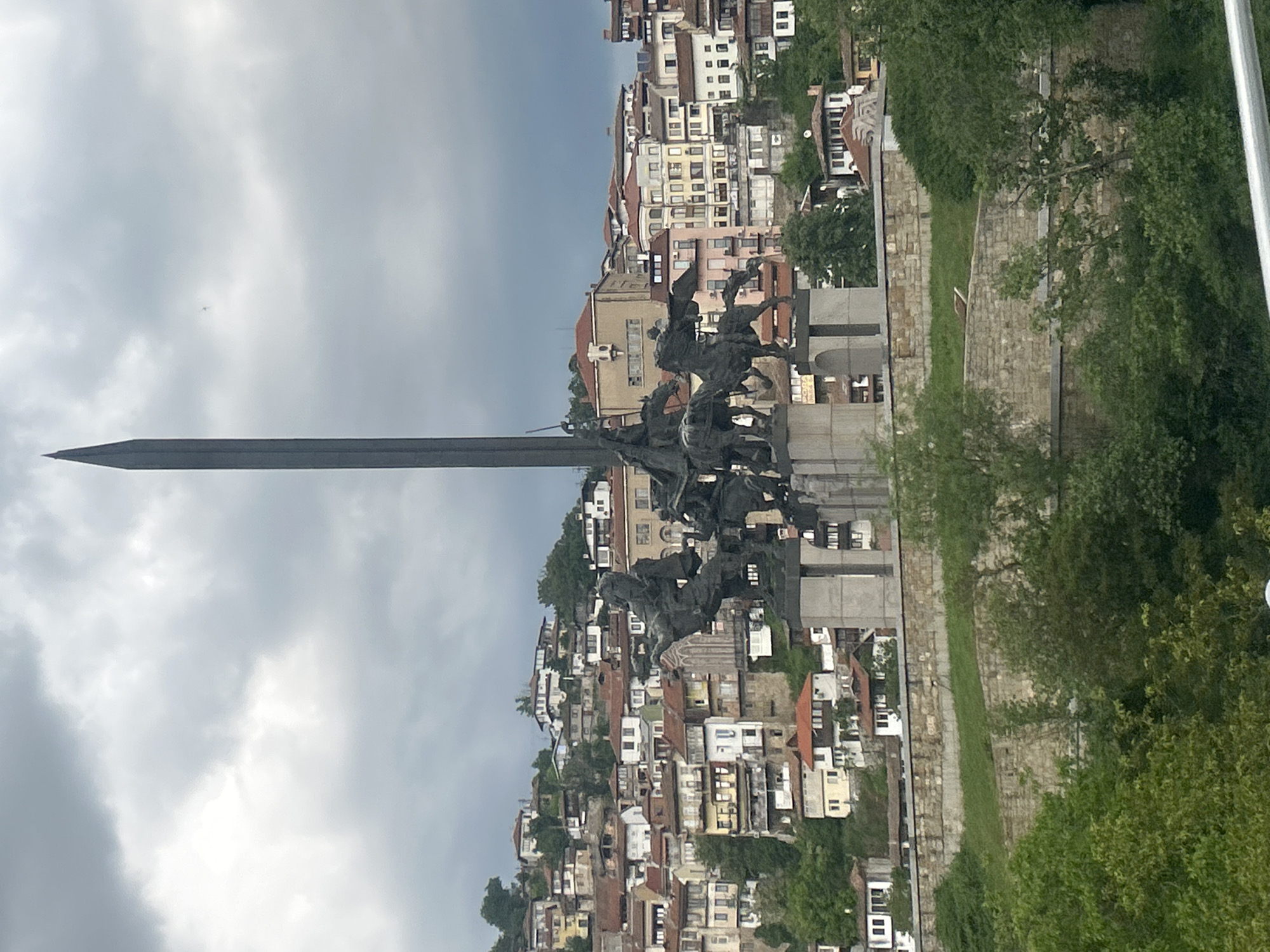 The image shows the Asenevtsi Monument in Veliko Tarnovo, Bulgaria, positioned against a backdrop of dense urban architecture. The monument depicts a group of riders on horseback and is surmounted by a large, vertically oriented sword. Buildings of varying heights and colors line the hillside behind the monument. The sky is overcast with gray clouds. Green trees and shrubbery are visible along the edges of the frame.