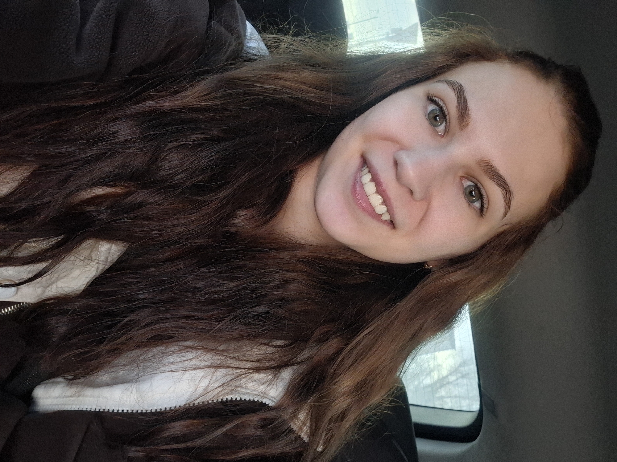 A selfie-style close-up image shows a young woman smiling broadly and looking directly at the camera, likely taken in Гаревая, Russia.
**Subjects:** The central subject is a woman with long, voluminous, wavy hair that is predominantly dark brown with lighter reddish-brown undertones and highlights. She has striking light green or hazel eyes and well-defined dark eyebrows. Her teeth are visible in her wide smile, and her complexion appears clear. She is wearing a dark-colored jacket, possibly made of fleece or a similar thick fabric, partially open to reveal a white inner layer or lining. A small, subtle golden earring is visible on her right earlobe.
**Activity or event taking place:** The woman is captured taking a self-portrait, posing with a cheerful and engaging expression.
**Notable details:** The setting appears to be indoors, most likely inside a vehicle such as a car, suggested by the dark interior trim and the shape of the window visible on the right side of the frame. Through this window, a bright, diffused light suggests it is daytime, with a blurry background showing what could be bare branches or distant structures. The woman's smile conveys happiness and approachability. There is no visible text in the image.