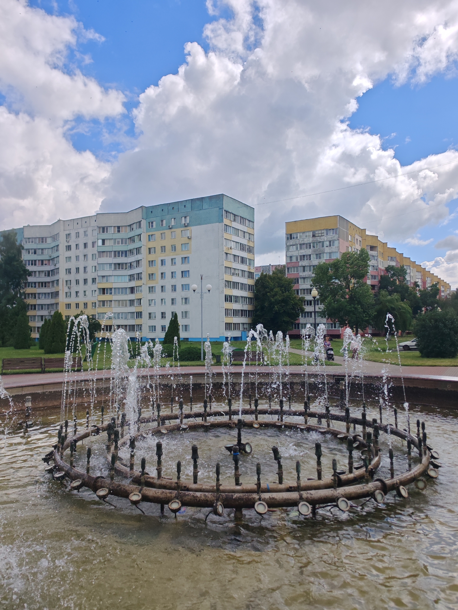 The image shows a circular fountain in the foreground with water jets spraying upwards. The fountain has a weathered metal structure with multiple nozzles. In the background, there are multi-story apartment buildings with beige, yellow, and light blue facades. Several trees are planted between the buildings and the fountain. A park bench is visible on the left side of the image, and a few people are walking in the background, one with a stroller. The sky is blue with scattered white clouds. This scene is located in the city of Rechytsa, Belarus.