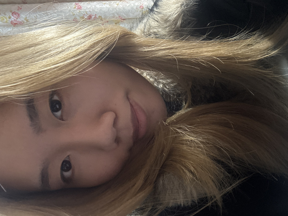 A close-up portrait depicts one East Asian female, appearing to be a young adult, with long, straight, blonde hair framing her face. She possesses dark eyes and a subtle, closed-lip smile, maintaining a direct gaze towards the viewer. The subject is wearing a dark outer garment, likely a jacket or coat, featuring a prominent faux fur collar visible around her neck and shoulders. The setting is indoors, with a background consisting of a light-colored patterned fabric, identifiable as a curtain or wallpaper, adorned with a floral design featuring pink and white blossoms. No discernible environmental markers are present to specify a location within Shapingba District, China.