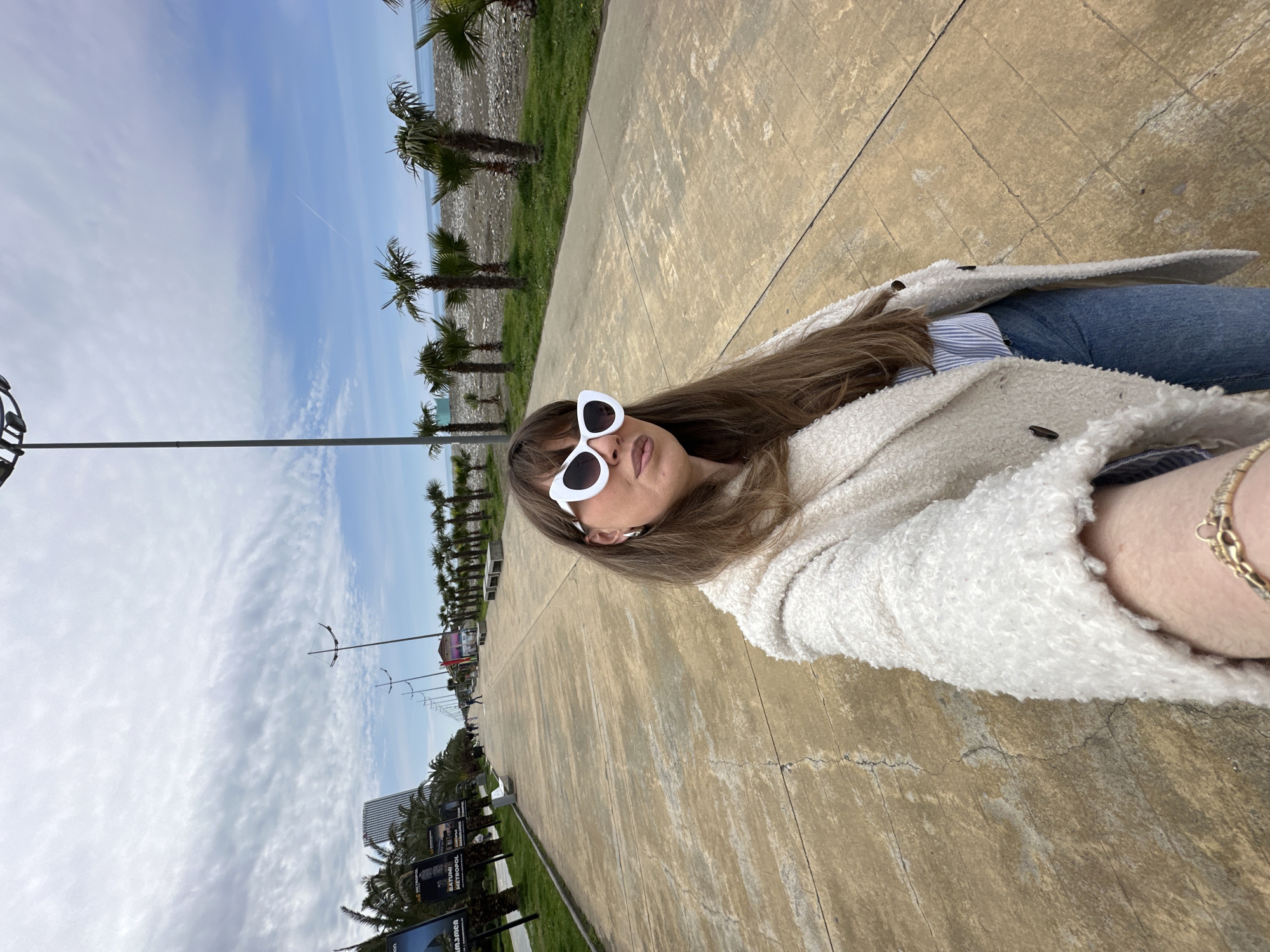 A medium shot, slightly angled, shows a woman wearing white-rimmed sunglasses and a white, textured coat, seated on a concrete surface. She is facing the camera. The background reveals a promenade lined with palm trees, running parallel to the coast. Street lamps are visible. On the left side of the frame, advertisements or billboards can be seen. The sky is partly cloudy with a hint of blue. The location is likely the Batumi Boulevard in Batumi, Georgia, a coastal promenade known for its palm trees and seaside views.