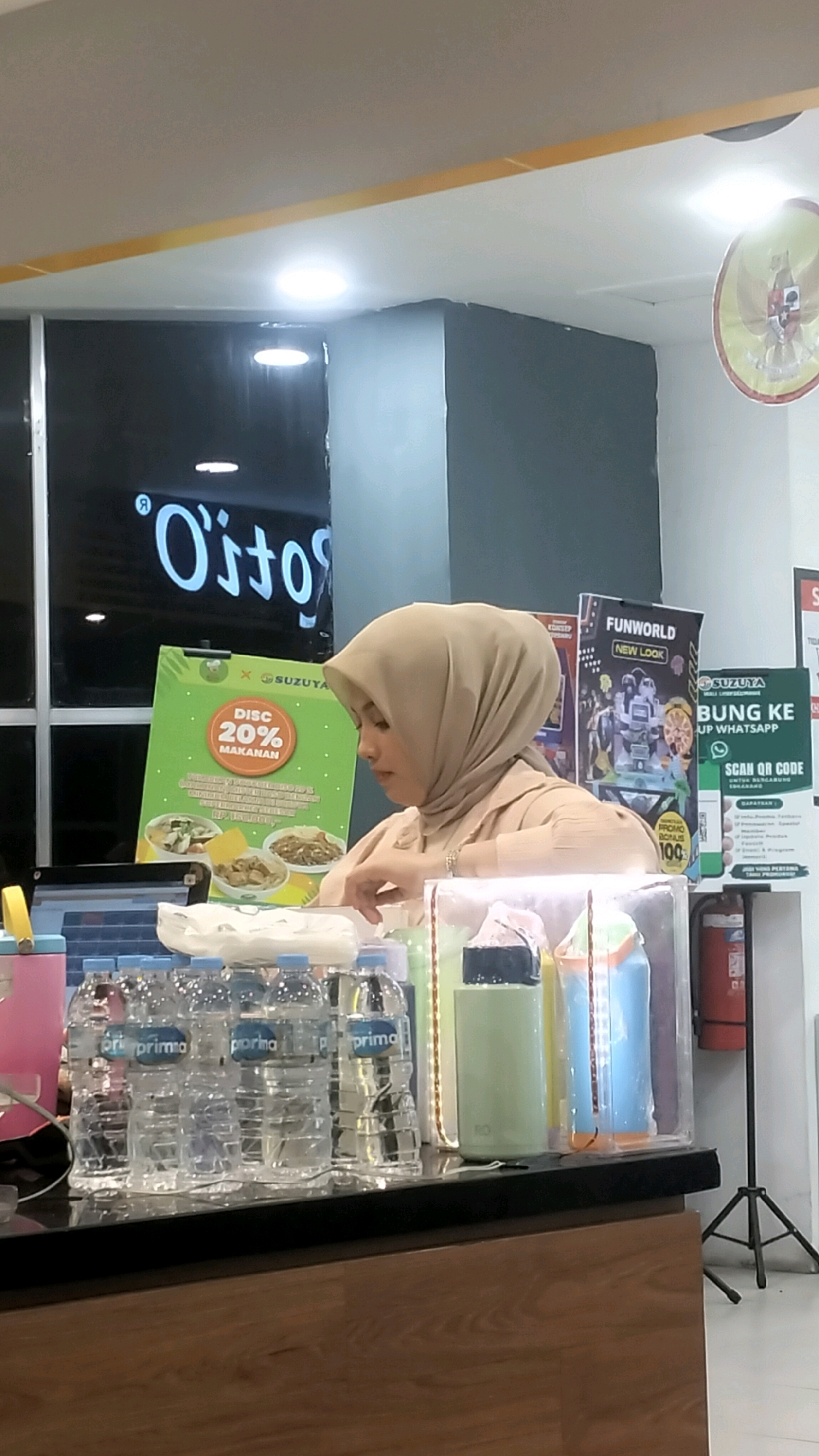 A woman wearing a light brown hijab and a matching shirt stands behind a counter in what appears to be a store or food establishment. She is looking down and to her right, with her hands near some items on the counter. In front of her, there are several bottles of water, some colorful containers, and a sign that says "DISC 20% MAKANAN" (20% Food Discount) with images of food. Behind her, there are posters, including one for "FUNWORLD NEW LOOK" and another with a QR code for WhatsApp. A large, dark sign with white lettering that appears to spell "O's to" is visible in the background. The environment is lit by overhead lights, and a circular Indonesian emblem is visible on the ceiling to the right. The floor appears to be tiled. The presence of promotional materials and a counter suggest a commercial setting, possibly a food stall or a retail store. The overall scene suggests a transactional environment where the woman is likely an employee.
