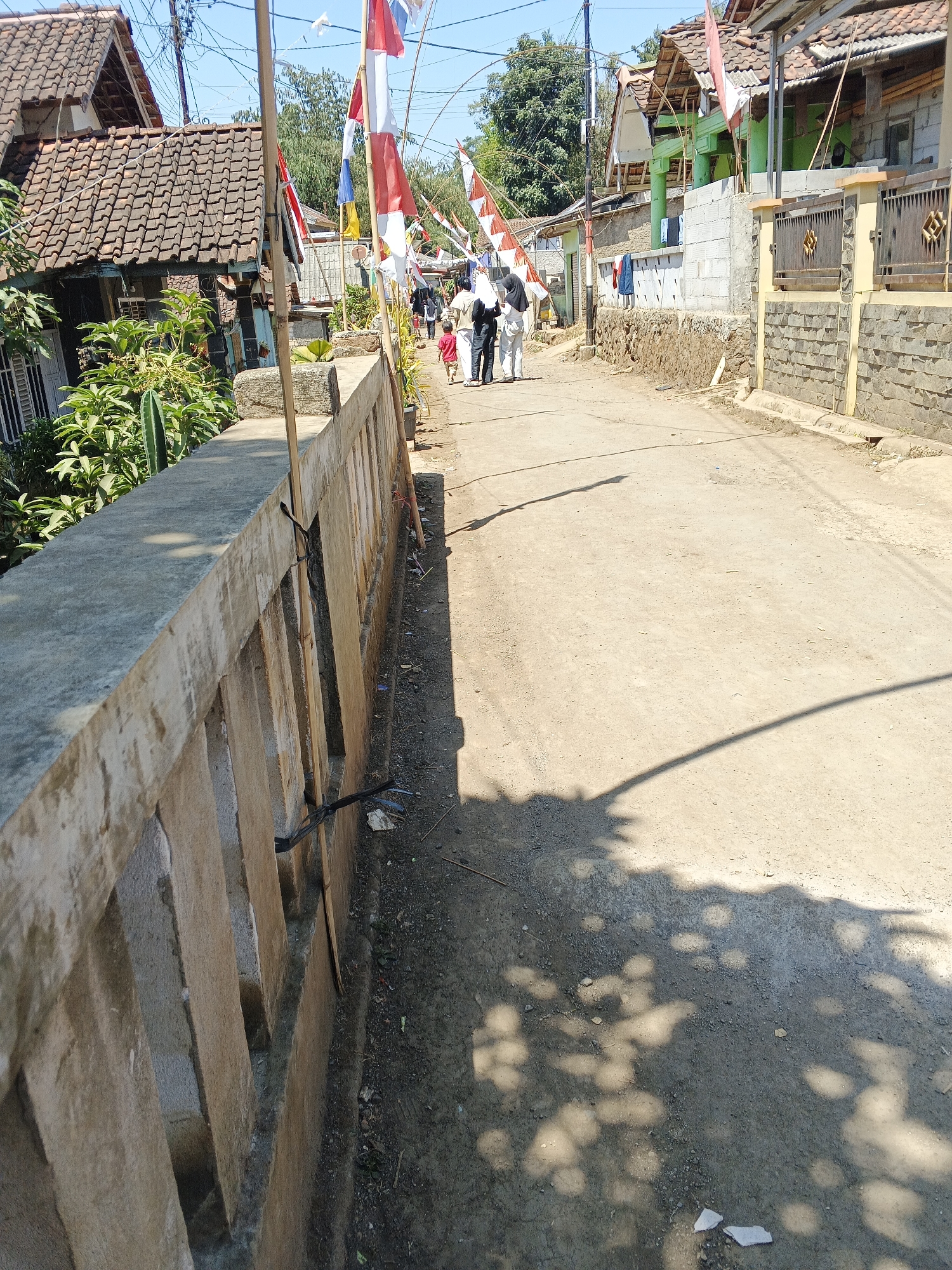 The image depicts a street scene in Sukaharja, Indonesia. The street is unpaved and appears to be dusty, with tire tracks visible. On the left side of the street, a low concrete wall runs along the edge. Beyond the wall, there are dense green plants and trees, with a house featuring a brown tiled roof behind them. Further down the street on the left, there are more houses with tiled roofs and what appear to be laundry hanging outside.
On the right side of the street, there are several people walking away from the camera. They are wearing modest clothing, with some individuals wearing headscarves. A young child in a red shirt is walking ahead of the adults. Various Indonesian flags are displayed on poles along the street, suggesting a celebration or national holiday. The houses on the right are varied in construction, with some featuring stone walls and others appearing to be under construction with exposed bricks. The overall atmosphere is sunny and dry.