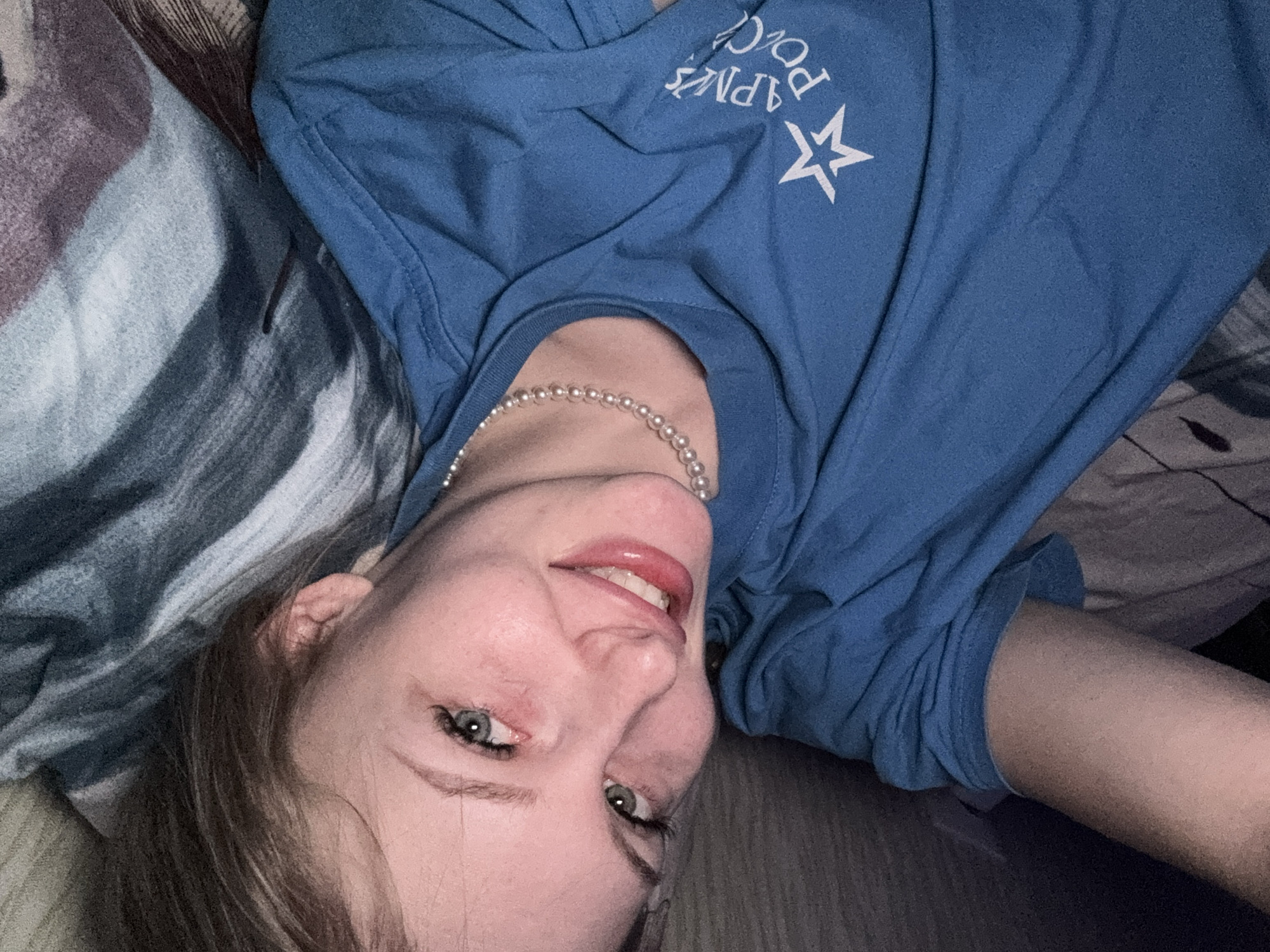 A young woman is depicted in a close-up, rotated portrait, likely a selfie, taken indoors. She is lying down, with her head positioned towards the bottom of the frame and her body extending upwards and to the right. She has fair skin, light hair, and green or blue eyes that meet the camera's gaze. Her lips are slightly parted in a gentle smile, revealing some teeth.
She is wearing a plain blue t-shirt adorned with white Cyrillic text and a white five-pointed star symbol. The visible text clearly reads "АРМИЯ РОССИИ" (Army of Russia). Around her neck, she wears a delicate pearl necklace. The background features patterned fabric in shades of grey, blue, brown, and white, possibly bedding or a blanket, creating a soft, textured setting. A darker, solid surface, likely wood, is visible towards the bottom and right edges of the frame. The lighting appears dim, suggesting an indoor environment. Given the text on her shirt and the provided context, the setting is in Кондратово, Russia. Her expression is calm and subtly joyful.