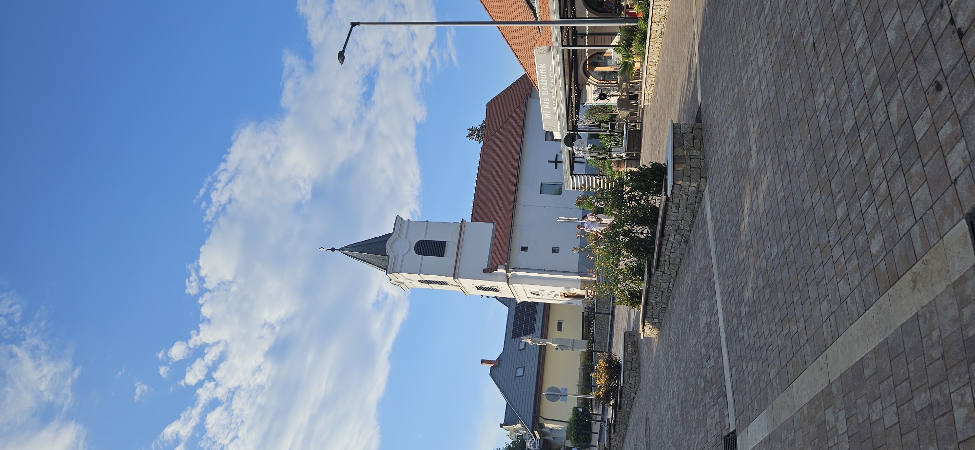 This is a photograph taken outdoors during the daytime, likely in the late afternoon given the shadows. The sky is a clear blue with scattered white clouds. The image features a prominent white church tower with a dark, pointed roof and a small cross at the apex. Adjacent to the church is a building with a red tiled roof, appearing to be a shop or restaurant, indicated by outdoor seating and plants. A sign visible on this building reads "Mi Kis Boltunk". The ground in the foreground is paved with a mosaic of light brown and grey rectangular tiles, suggesting a public area like a square or street. There are some trees and bushes visible, adding greenery to the scene. The overall atmosphere appears peaceful and sunny.