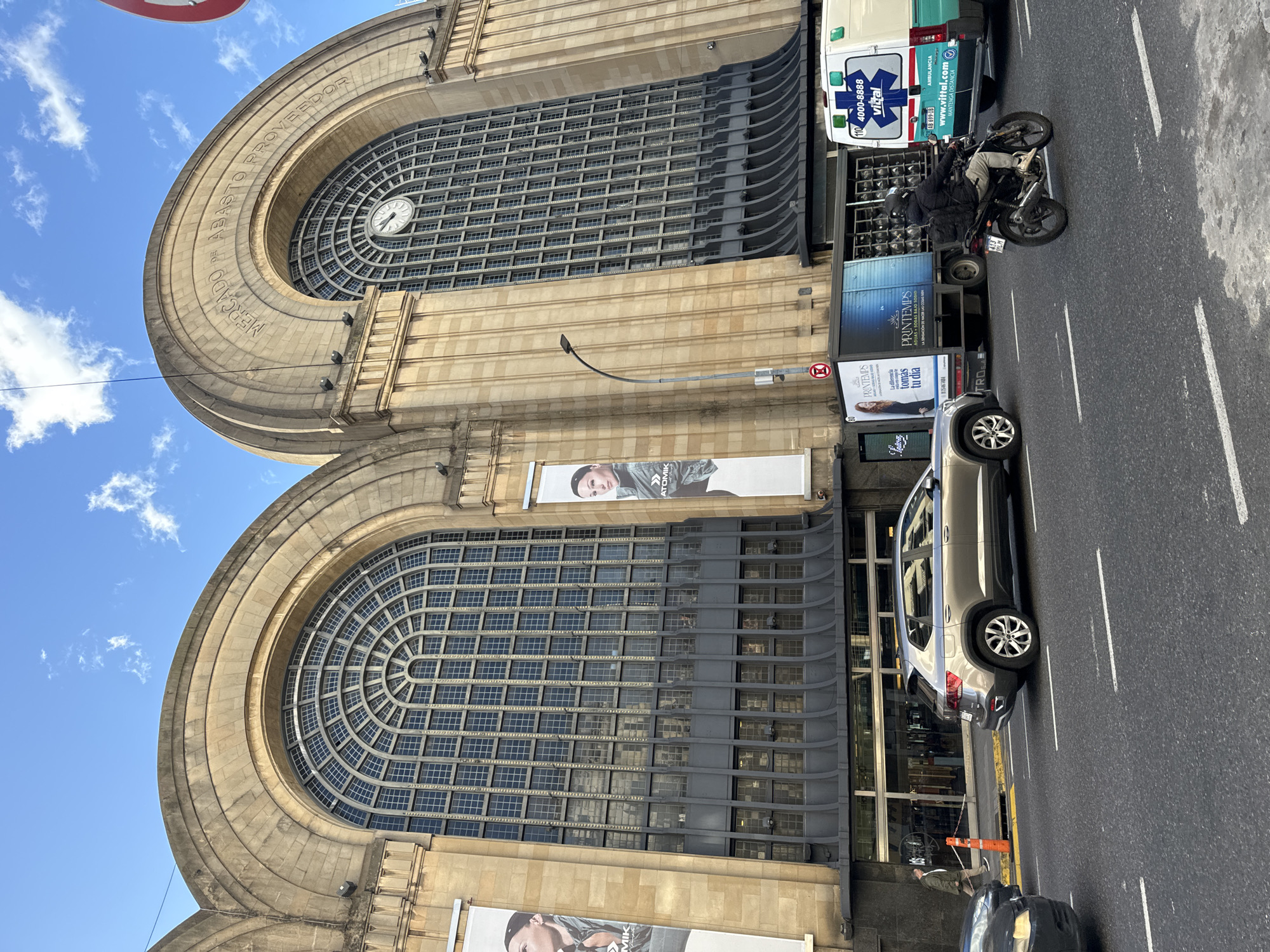 The image shows a partial view of El Abasto, a shopping mall in Buenos Aires, Argentina. The facade of the building, a former wholesale produce market, is visible, with its distinctive arched windows and stone inscription "MERCADO DE ABASTO PROVEEDOR". Below the windows, there is an advertising poster featuring a model. The street in front of the building contains a silver SUV, a delivery truck, an ambulance, and a motorcycle parked on the side of the road. Additionally, a person can be seen walking on the sidewalk.