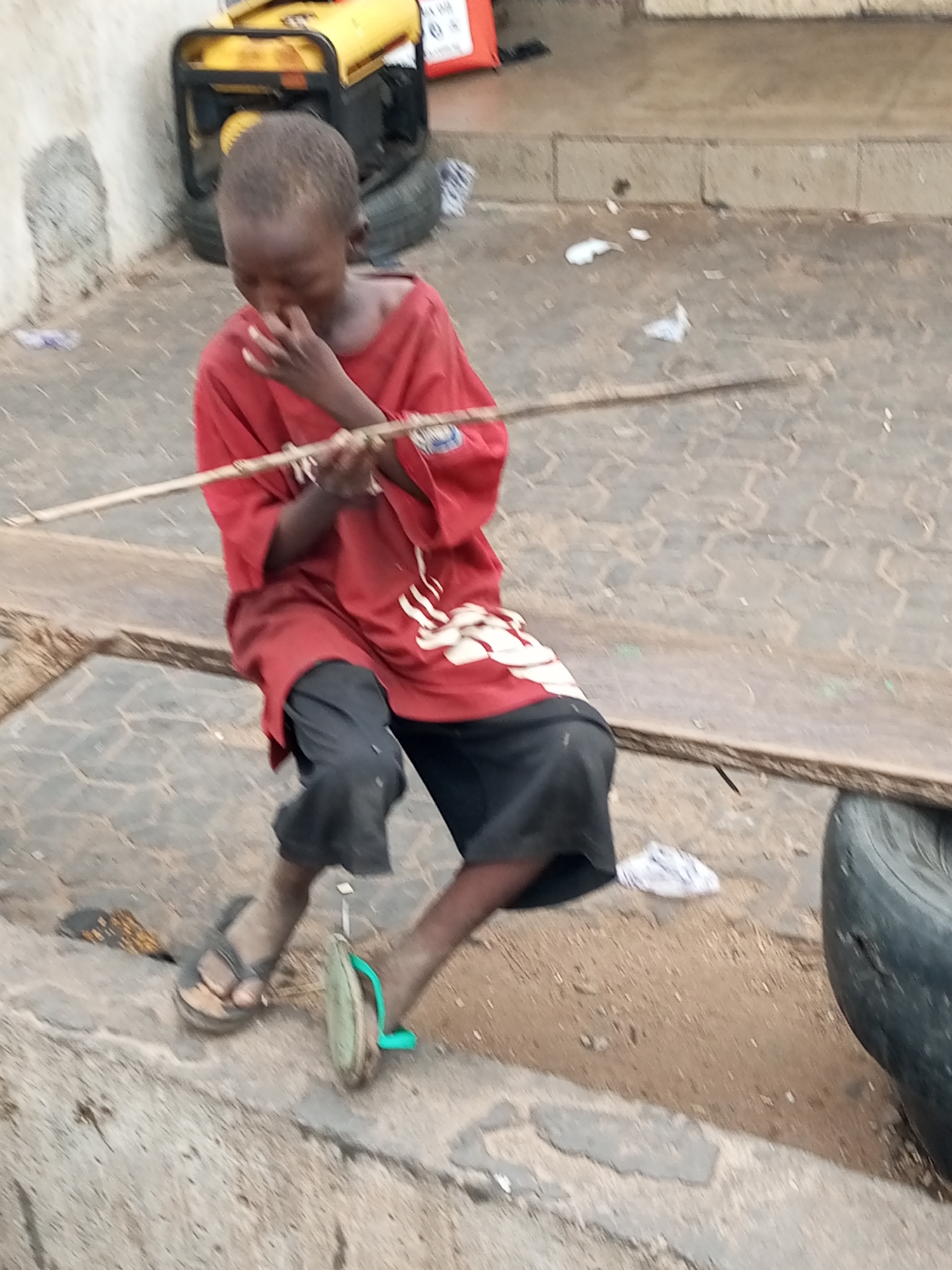 A young boy, appearing to be of primary school age, sits on a raised concrete curb. He is wearing a red t-shirt with a white graphic design and dark shorts. His left hand is raised to his face, and he is holding a thin wooden stick horizontally with both hands. He is wearing a dark sandal on his left foot and a light green flip-flop on his right foot. The environment appears to be an outdoor urban setting. In the background, to the left of the boy, is a yellow and black generator resting on tires. The ground is paved with interlocking blocks, and there is some dirt and debris scattered around. The scene suggests a casual, possibly impoverished, setting within the city of Gombe, Nigeria.