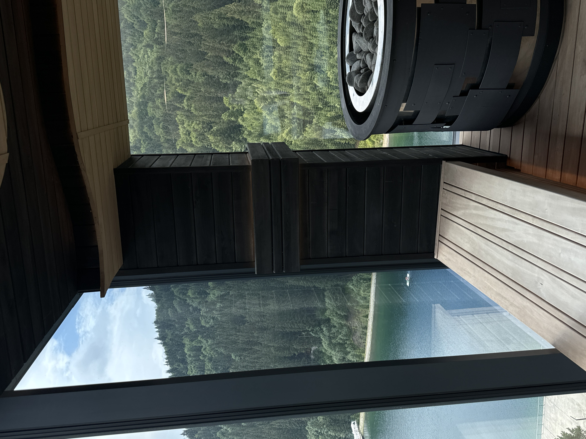 The image captures an interior view of a modern sauna room overlooking a scenic natural landscape. The room features dark wooden paneling on the walls and ceiling, contrasting with a light-colored wooden bench. To the right, a black metal cylindrical sauna heater filled with dark stones is visible. Large floor-to-ceiling windows offer a panoramic view of a serene lake and a densely forested mountain range under a cloudy sky. In the distance, a small white paddle boat with two occupants is visible on the lake. The location is identified as Polianytsia, Ukraine, a resort village known for its natural beauty and proximity to the Carpathian Mountains.