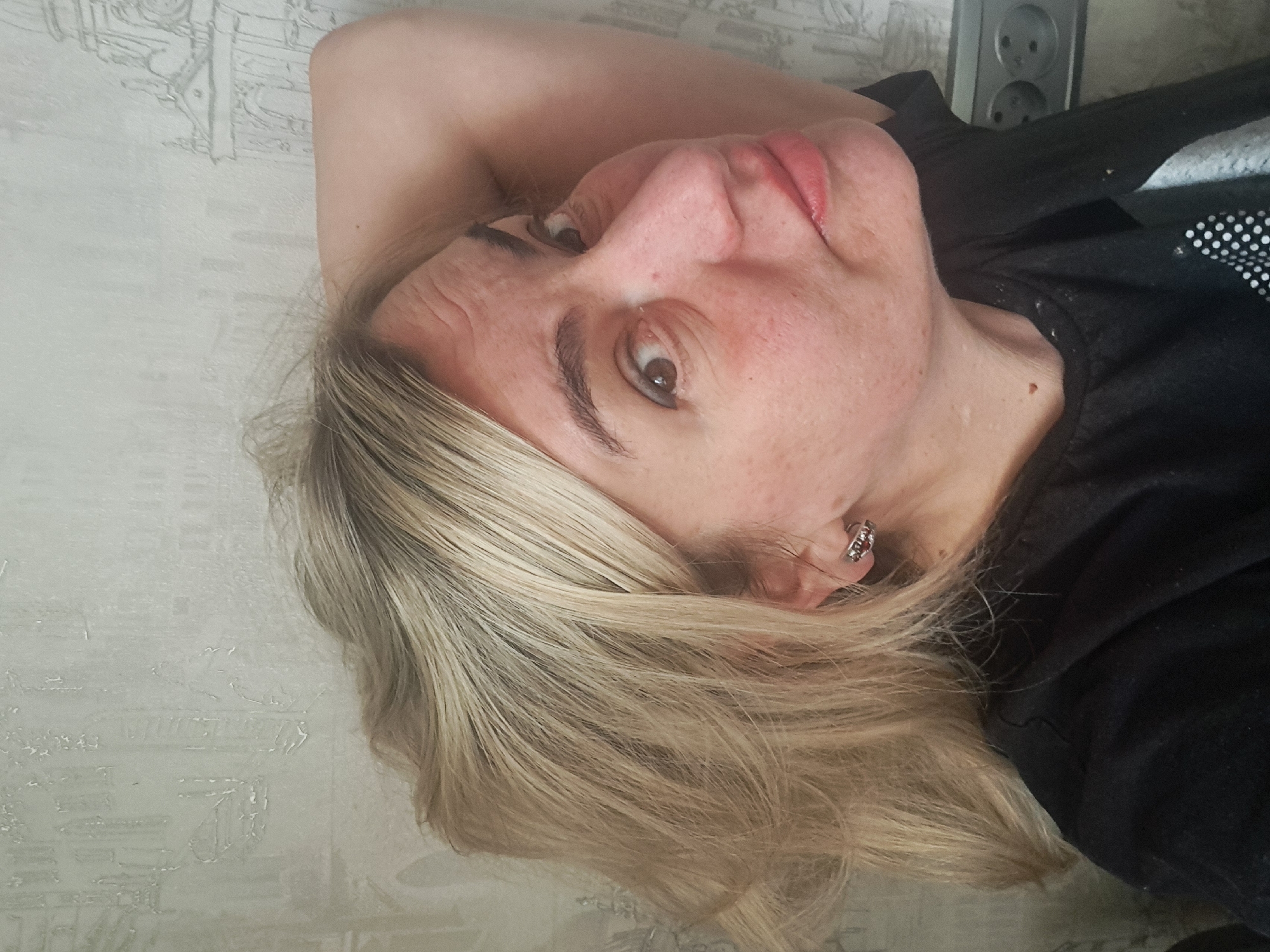 The image depicts a female individual in an indoor setting within Elektrostal, Russia. The subject is a woman with blonde hair, visible brown roots, and light skin, captured in a close-up, chest-up view. Her head is tilted slightly to her left, and her gaze is directed towards the upper left from the viewer's perspective, with her right eye clearly visible. Her right arm is raised, with the elbow visible behind her head. She is wearing a black t-shirt or long-sleeved top, partially obscuring a white spotted pattern on the garment. A small, silver-colored hoop earring is present on her left earlobe. The background consists of light-colored wallpaper featuring a faint, repeating pattern resembling stylized architectural structures. In the lower right corner of the frame, a gray electrical outlet with two sockets is mounted on the wall.