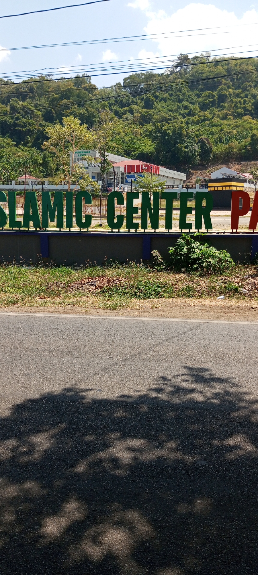 The image shows a sign in front of the Islamic Center in Lempa, Indonesia. The sign has large, green, three-dimensional letters spelling out "ISLAMIC CENTER PA". The letters are mounted on a dark grey wall with blue trim. Behind the sign, there is a building with a white facade and red roofing, identified as "PUSKESMAS LEMPA" and "UGD". Further in the background, lush green hills covered in trees rise up under a bright blue sky with some power lines visible. In the foreground, there is a paved road with shadows from trees cast upon it, and a grassy area with some dry leaves and sparse vegetation along the edge of the road.