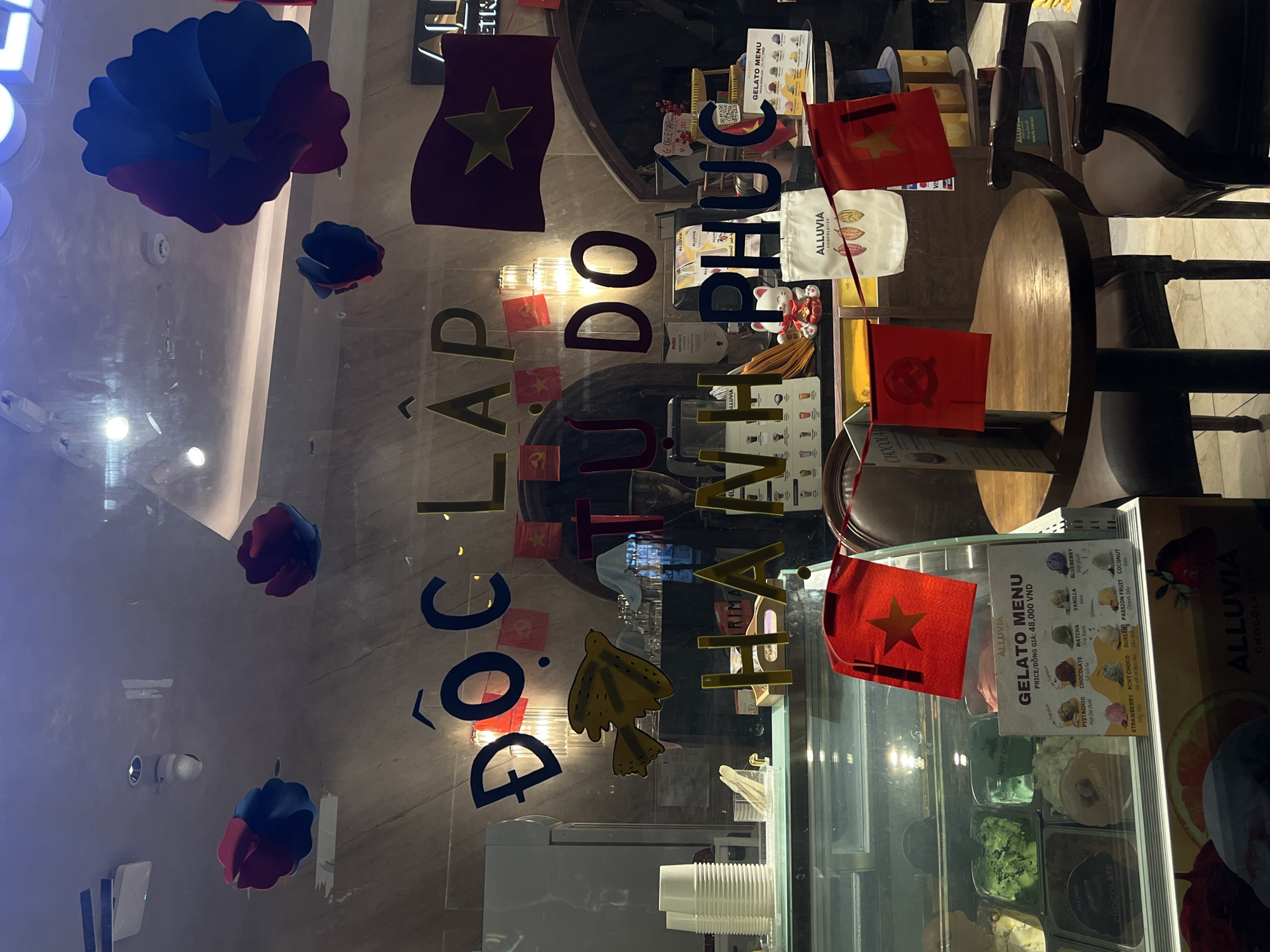 Here is a description of the image:
The image is an interior shot, likely taken from inside a store or cafe called "Alluvia," specializing in gelato and chocolates. The storefront window is decorated with Vietnamese flags and floral decorations, with lettering on the glass spelling out "ĐỘC LẬP TỰ DO HẠNH PHÚC," which translates to "Independence, Freedom, Happiness." This suggests a celebration of Vietnam's national day.
The interior displays a gelato menu and freezers filled with different gelato flavors. A wooden table and chair are visible in the background, suggesting a seating area. The view through the window shows reflections and glimpses of the outside environment, including street lights and a hint of urban activity, although specific location details are not available. The presence of the Alluvia branding on the gelato menu and chocolate packaging indicates that the focus is on the store's products and its festive decoration.