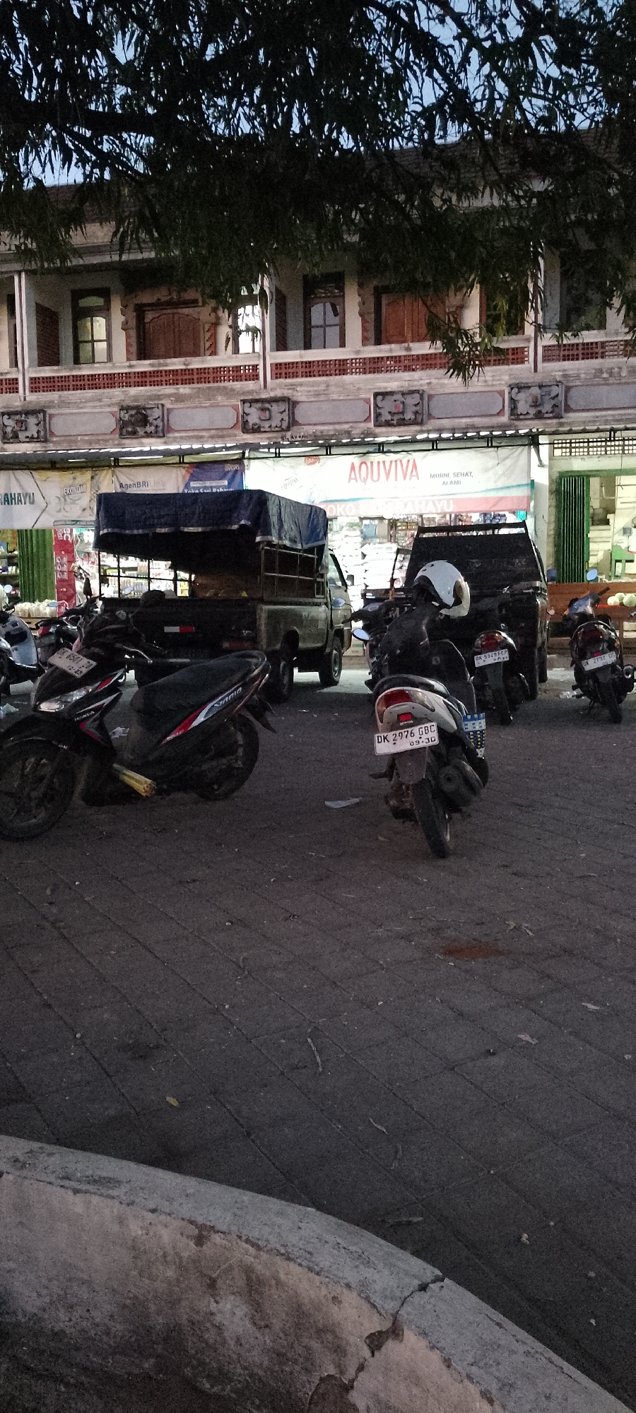 The image depicts a street scene in Pekutatan, Indonesia, with several motorcycles parked in the foreground. Behind the motorcycles, a covered pickup truck is visible, positioned in front of a building with a banner that reads "AQUVIVA." The building appears to be a commercial establishment, possibly a shop or market, with signage and open doorways. The street is paved with interlocking concrete blocks. Overhead, tree branches obscure part of the sky and the upper portion of the building. The overall atmosphere suggests an outdoor marketplace or commercial area.