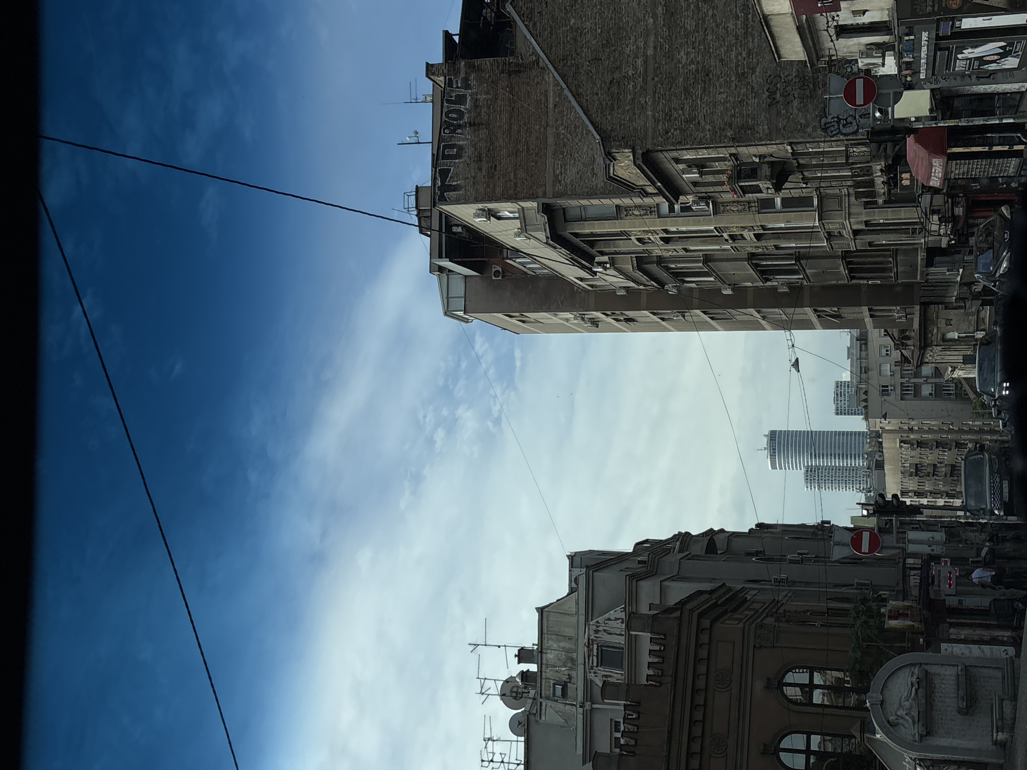 The image shows a street scene in Belgrade, Serbia, viewed from a low angle, possibly from a car. Buildings line both sides of the street, with varying architectural styles, indicating a mix of old and newer structures. One building has graffiti on its upper portion. A modern high-rise building is visible in the distance between the buildings. Traffic signs are present, one is a no entry sign, indicating traffic regulations on the street. Wires are strung across the sky, contrasting with the blue sky and clouds. The scene is densely urban, with a mixture of residential and commercial buildings, suggesting a busy city environment.