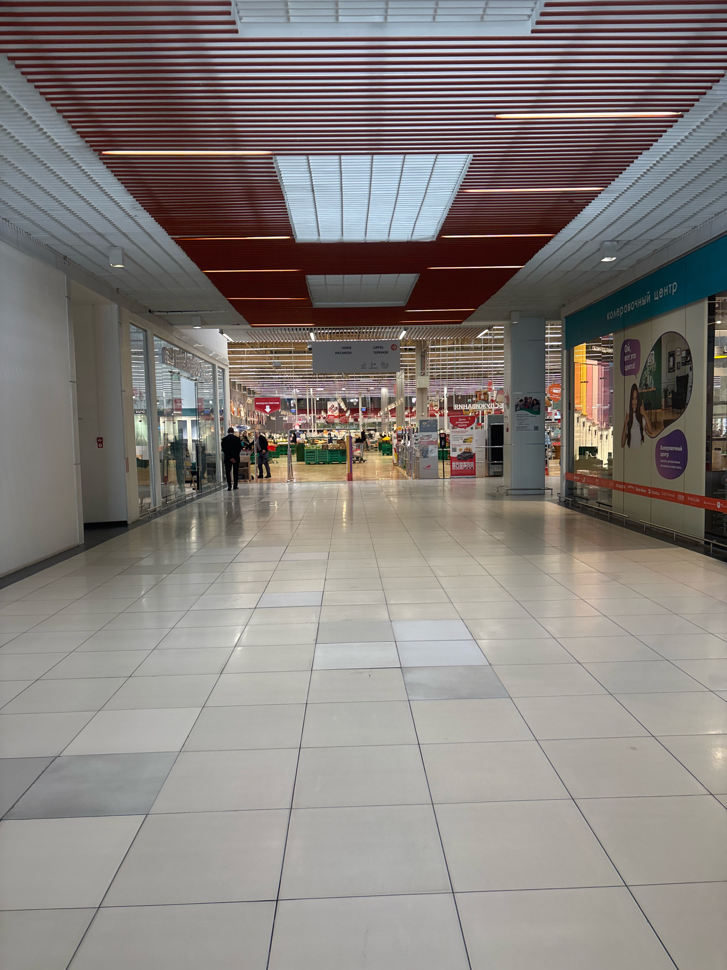 The image depicts a bright, modern interior hallway of a large retail establishment, possibly a supermarket or a shopping mall, with clean, light-colored tiled flooring. The hallway is wide and extends into the distance, lined with storefronts on either side. The ceiling features a complex design with red and white horizontal slatted panels and recessed lighting. On the right side, a storefront with blue signage that reads "КОЛЕРОВочный центр" (Color Center) is visible. Several people are present in the hallway, some walking and others standing near store entrances. In the mid-ground, a man in a black suit is walking away from the camera, and further ahead, two individuals are standing near a store entrance. In the background, a large retail area with aisles of products and more shoppers is visible. The overall impression is one of a busy, commercial environment. The location is unknown.