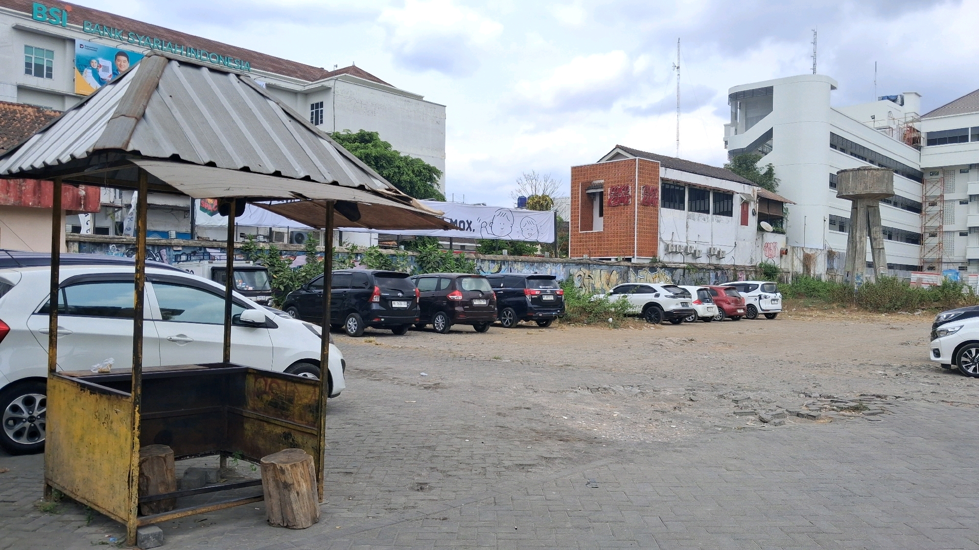 The image captures a parking lot in front of a building in Yogyakarta, Indonesia. A small, weathered food stall with a corrugated metal roof occupies the foreground, to the left. Behind it, several parked cars are visible, including a white hatchback, several black SUVs, and a maroon minivan. The building in the background is a multi-story structure with a white facade and dark windows. A large banner with graphics and text is displayed on the building. Further in the background, a water tower and other buildings are visible, suggesting an urban or semi-urban setting. The ground is paved with interlocking concrete blocks in the foreground and a rougher, dirt-and-paved surface in the parking area. The sky is overcast. There are no people visible in the image.