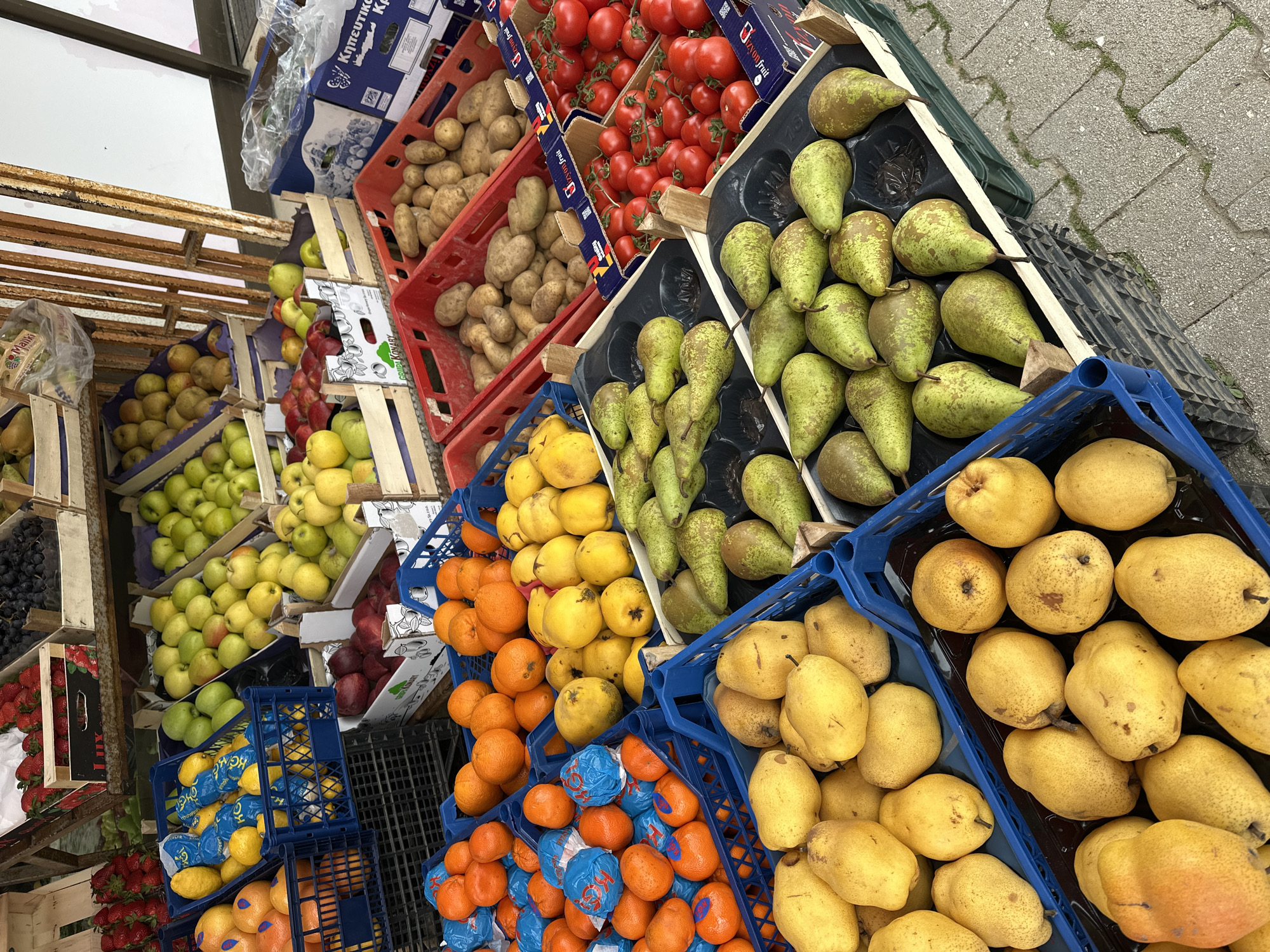 The image shows a street-side produce stand in Šušanj, Montenegro. Several crates are stacked with fruits and vegetables. Red crates contain potatoes and tomatoes. There are pears with green, yellow, and brown skin and lemons in blue-wrapped packaging. The stand also displays apples, oranges, and tangerines in blue-wrapped packaging. The crates are arranged on the sidewalk, which is gray and has irregular cracks. The stand is partially shaded by a structure above.