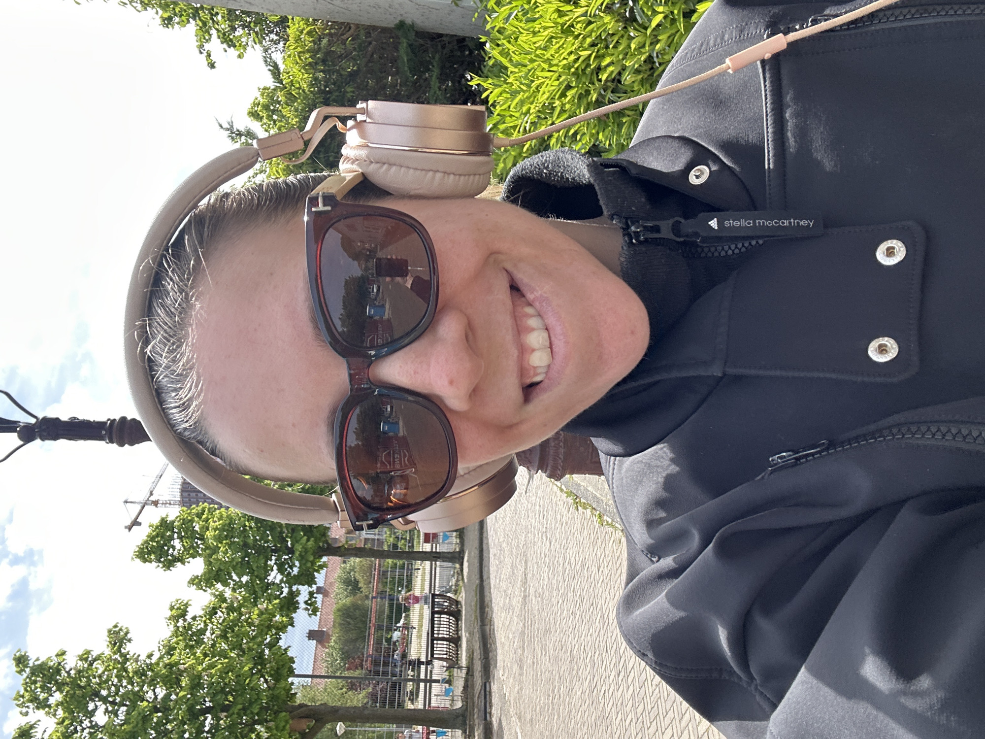 A fair-skinned person with sunglasses and headphones stands on a paved sidewalk. The person is wearing a black jacket with a Stella McCartney logo visible. In the background, there are trees, a fence, and a building. A construction crane is also visible in the distance. The photo was taken in Amsterdam, Netherlands.