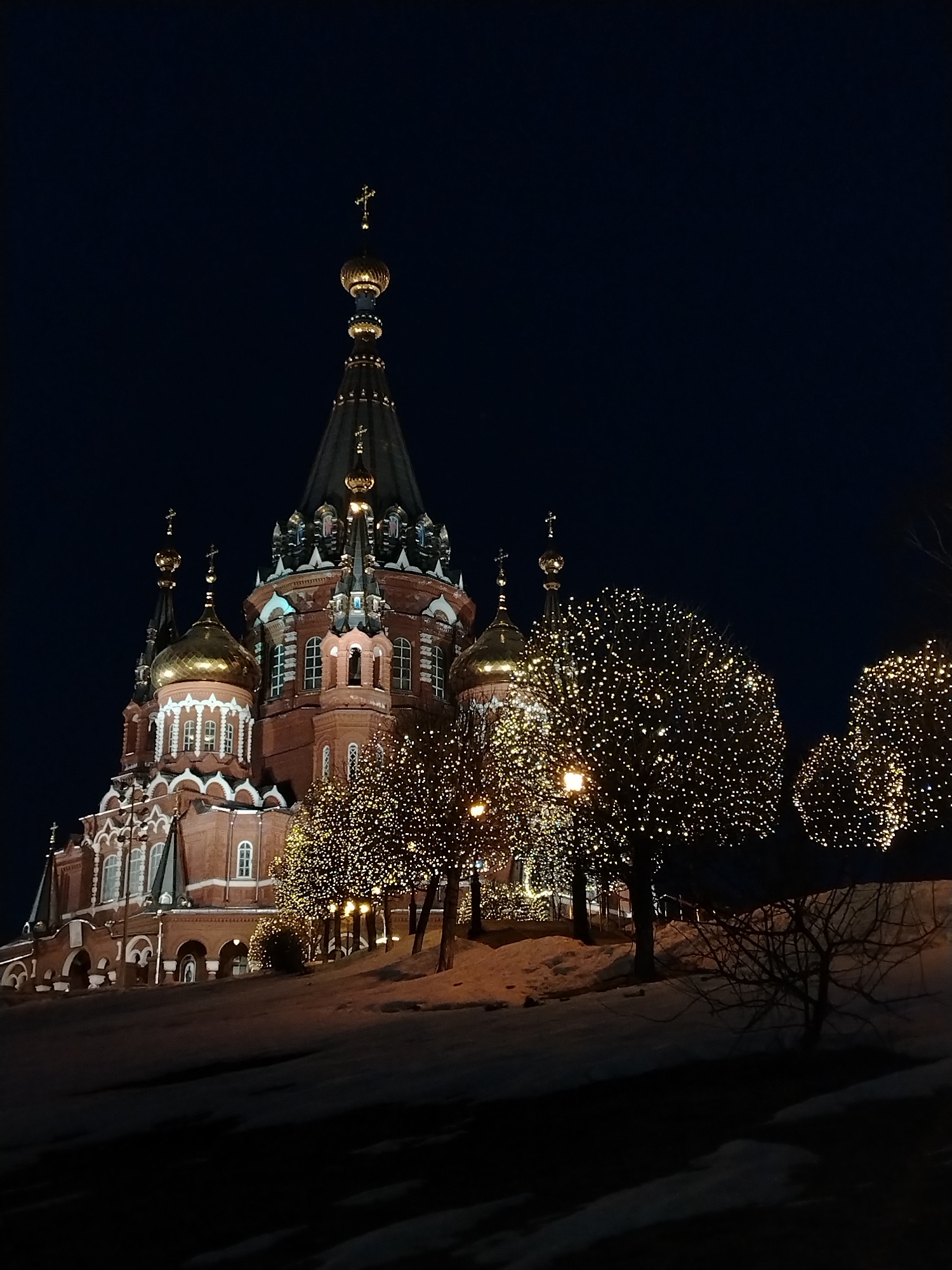 The image depicts the Cathedral of Alexander Nevsky in Izhevsk, Russia, at night. The cathedral, constructed of red brick with white detailing, has multiple golden domes and a towering central spire topped with a golden cross. The building is illuminated with lights that accentuate its architectural features.
In the foreground, several trees are decorated with strings of lights, creating a warm, twinkling effect. The ground is covered with snow. The overall atmosphere is festive and serene, with the dark sky providing a contrasting backdrop to the brightly lit cathedral and trees. No people are visible in the frame.