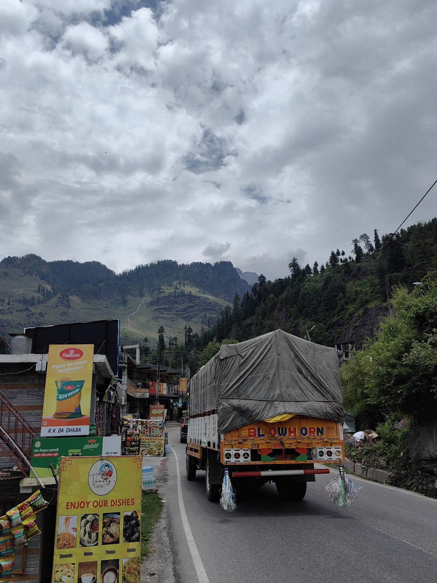 The image captures a view of a street in Palchan, India, under a cloudy sky. In the foreground, a large truck covered with a grey tarp is driving on the right side of the road. The truck has colorful lettering on its rear that reads "BLOW HORN". On the left side of the street, there are several signs and small shops. One prominent yellow sign displays "Tamang's Taste" and lists various dishes like Chowmin, Momo, Thukpa, and Gyuma. Another sign advertises "Haldiram's" snacks, including "Aloo Bhujia". Further down the road, more shops are visible, and bottles of water are stacked by the roadside. Behind the shops and road, a lush green mountain range covered with trees rises steeply. The overall scene depicts a lively roadside environment in a mountainous region.