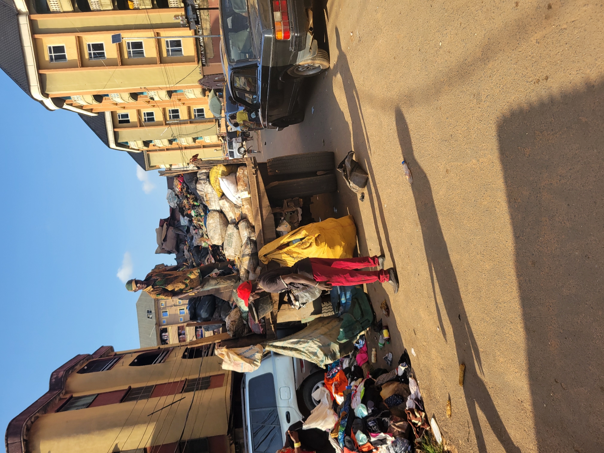The image depicts a street scene in Onitsha, Nigeria, characterized by a large quantity of refuse. In the foreground, a man wearing a red cap and red trousers stands near a pile of discarded clothing and other items. Behind him, a flatbed truck is heavily laden with trash bags and loose refuse, some of which appears to be clothing and textiles. Several large, yellow trash bags are prominent on the truck and near the man.
The environment suggests an urban setting with multi-story buildings lining the street. One building on the left is painted yellow and brown, while buildings on the right are a lighter yellow with black roofs. A silver SUV is parked to the left of the truck. Further down the street, a black car is visible. The ground is a paved asphalt surface, with shadows from the objects and people cast across it, indicating daylight. The overall scene suggests a collection point or a temporary storage area for waste in a busy urban area.