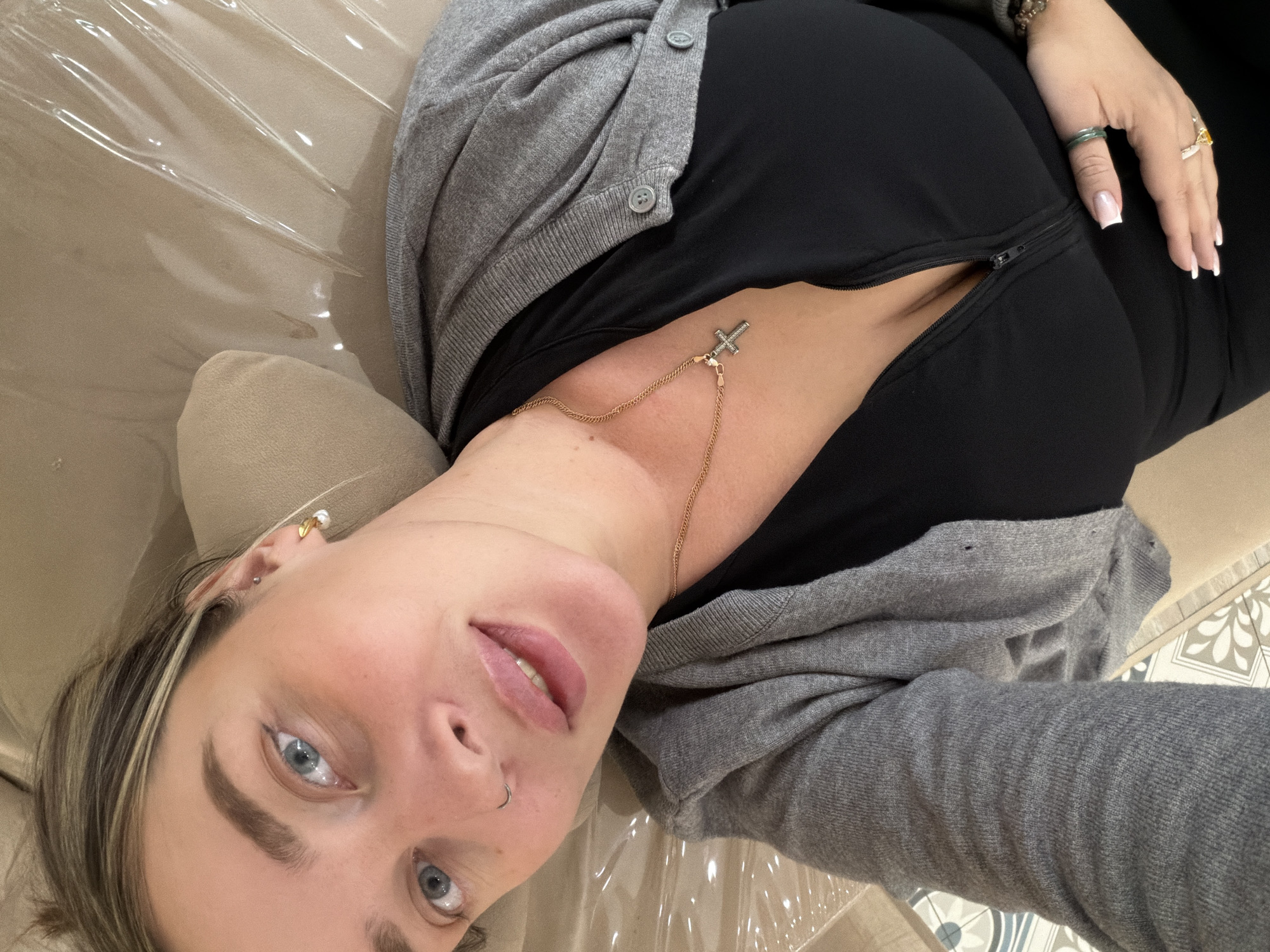 A woman lies on a light-colored surface, likely a couch or treatment bed, covered with a clear plastic sheet, looking directly into the camera. Her head is tilted to the left, and her body extends towards the upper right of the frame. She has light skin, blue eyes, and light brown hair with blonde highlights. A small hoop piercing is visible in her left nostril, and she wears a gold-colored earring with a pearl in her right ear.
She is dressed in a black, form-fitting top with a partially unzipped zipper revealing a glimpse of skin on her side. Over this, she wears a gray knitted cardigan or sweater with visible buttons. A delicate gold-toned chain with a small, sparkling cross pendant rests on her chest. Her right hand, adorned with several rings including a green band and a large yellow-stoned ring, and a beaded bracelet, is visible towards the upper right, displaying French-tipped nails.
The background includes a hint of a decorative tiled floor with geometric and floral patterns. The setting appears to be indoors, possibly in Khimki, Russia. The lighting is soft and even. No visible text is present.