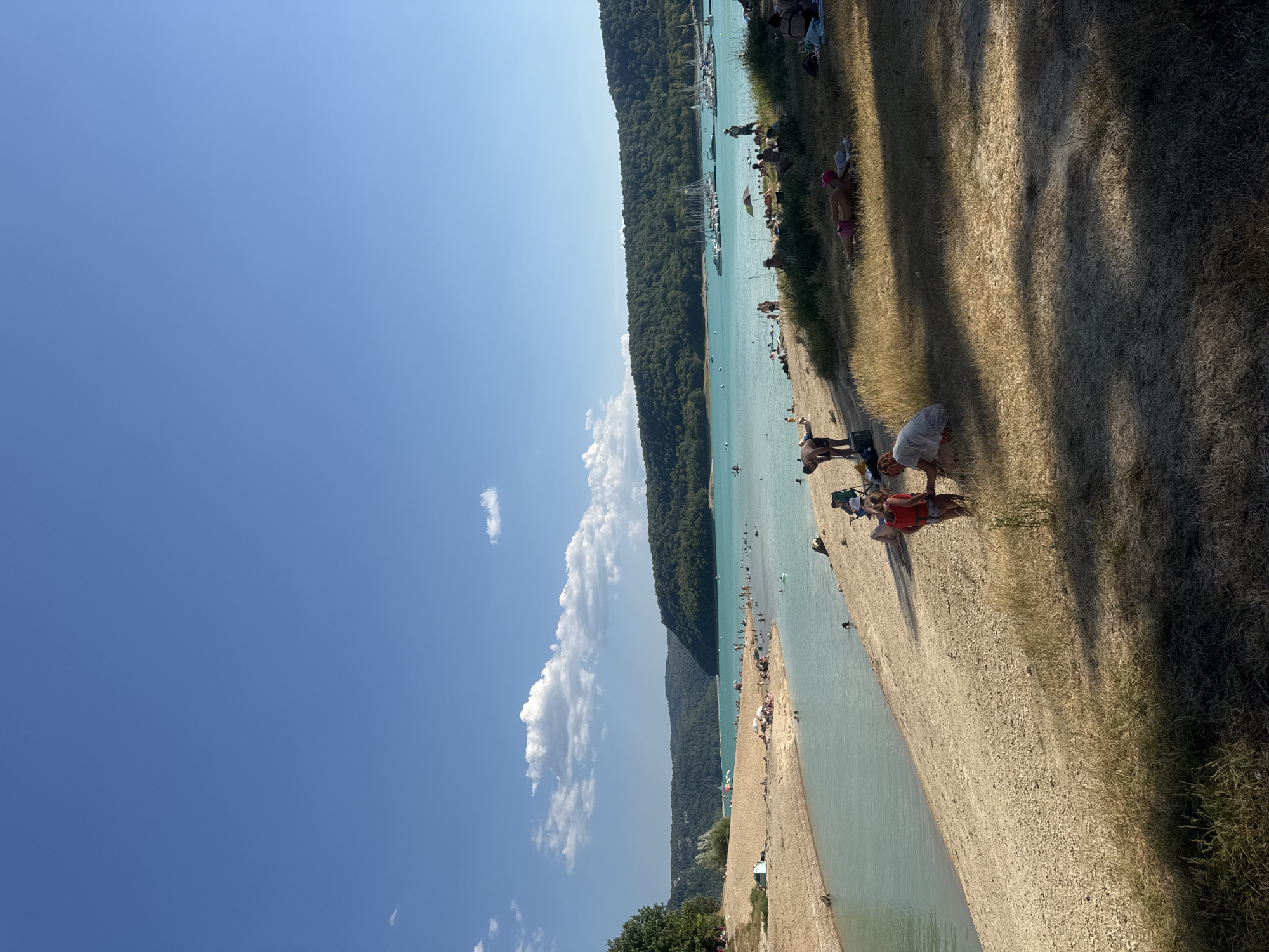 This image depicts a sunny day at a beach on a lake, likely Lac de Vouglans, near the village of Maisod, France. The scene is filled with people enjoying various beach activities. In the foreground, several individuals are sunbathing on the grassy shore, with one person in a pink hat and another in a white cover-up. Further back, on the sandy beach, a child wearing a red life vest is interacting with another person. The lake itself is populated with swimmers and people wading. A small inlet with shallow, clear turquoise water separates the sandy beach from a wider body of water. In the middle ground, a line of sailboats are docked at a marina. The background is dominated by a lush, tree-covered hillside under a clear blue sky with scattered white clouds.