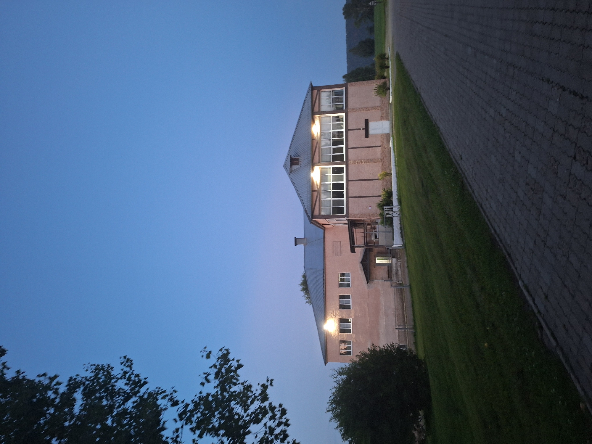 The image depicts an exterior view of a building at dusk in Атамановка, Russia. The building, with a light pink facade and a grey metal roof, features multiple windows and an entrance with an awning. The upper section of the building has large, multi-pane windows, suggesting a large interior space. The ground in front of the building is a manicured lawn that slopes gently downward, with a paved walkway to the right. The sky is a clear, deep blue, transitioning to a lighter shade near the horizon, indicating the twilight hours. A large tree with dark foliage partially obscures the upper left corner of the frame. The scene is devoid of any visible people or animals.
