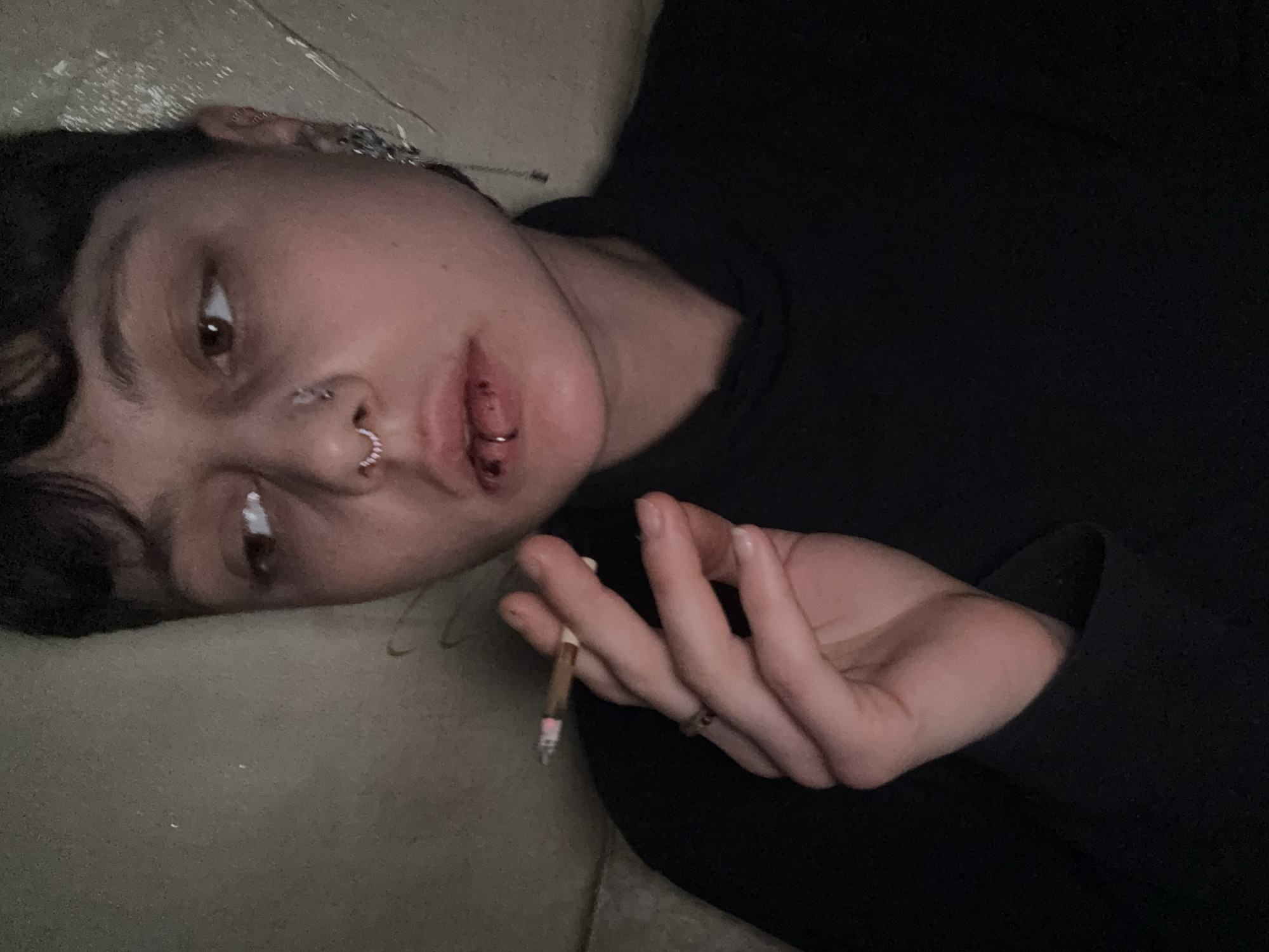 The image features a young individual lying horizontally on a light-colored, possibly beige or grey, surface. The person has short, dark hair and is looking slightly upwards and to the right with brown eyes, displaying a neutral, relaxed, or introspective expression. They are wearing a dark, likely black, long-sleeved top.
Notable facial features include several piercings: a silver circular barbell in their septum, a similar circular barbell piercing through their lower lip (labret piercing) with a reddish tint around it suggesting it might be new or irritated, and multiple earrings in their visible left ear, including hoops and a chain detail.
In their right hand, held between the index and middle fingers, is a lit cigarette or joint, with a visible glowing tip and ash. A ring is also worn on one of the fingers of this hand. The scene is dimly lit, indicating an indoor setting at night or under low light conditions. The overall atmosphere is quiet and intimate. The location context provided for this media file is Moscow, Russia.