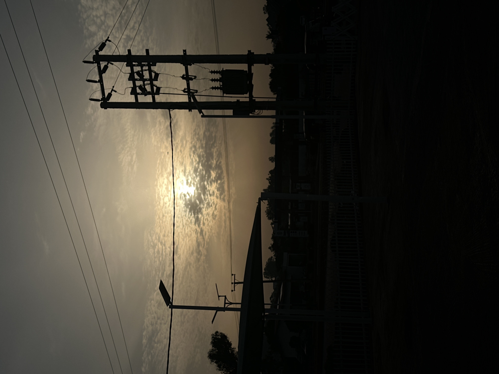 The image captures a silhouette view of utility poles and power lines against a bright, hazy sky. The sun is partially obscured by clouds, casting a glow around them. A streetlight and a partially visible structure with a fence are also silhouetted. The foreground fades into darkness. While the prompt requested a location in Bukuru, Nigeria, there is nothing in the image to specifically confirm this location.