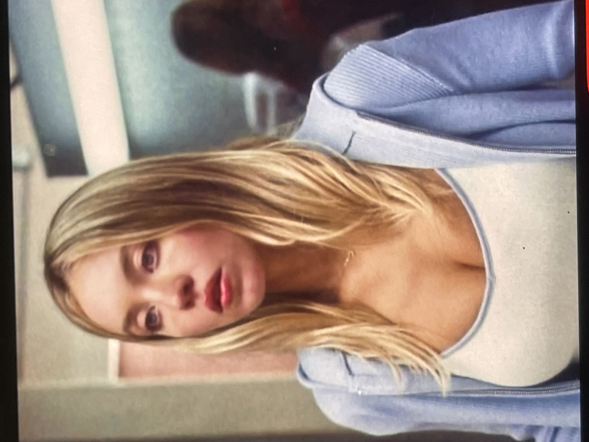 The image features a young woman with long, straight blonde hair and fair skin. She is wearing a light blue hooded jacket or sweatshirt, partially unzipped, revealing a light-colored (possibly white or off-white) scoop-neck top underneath. A thin, delicate necklace is visible around her neck. Her eyes appear to have a reddish or purplish tint, possibly from makeup or lighting, and her lips are parted, showing a red lip color. Her expression is neutral, looking slightly off-camera.
The background is out of focus, suggesting an indoor setting with muted tones, possibly walls or a simple room. There's an indistinct dark shape in the upper background, which might be another person or an object. The lighting is soft and diffused. The overall image appears to be a still frame, possibly from a video, and is rotated on its side. No specific activity is discernible beyond her looking. There is no visible text. The image does not contain any visual cues to confirm the specified location of Poltava, Ukraine.