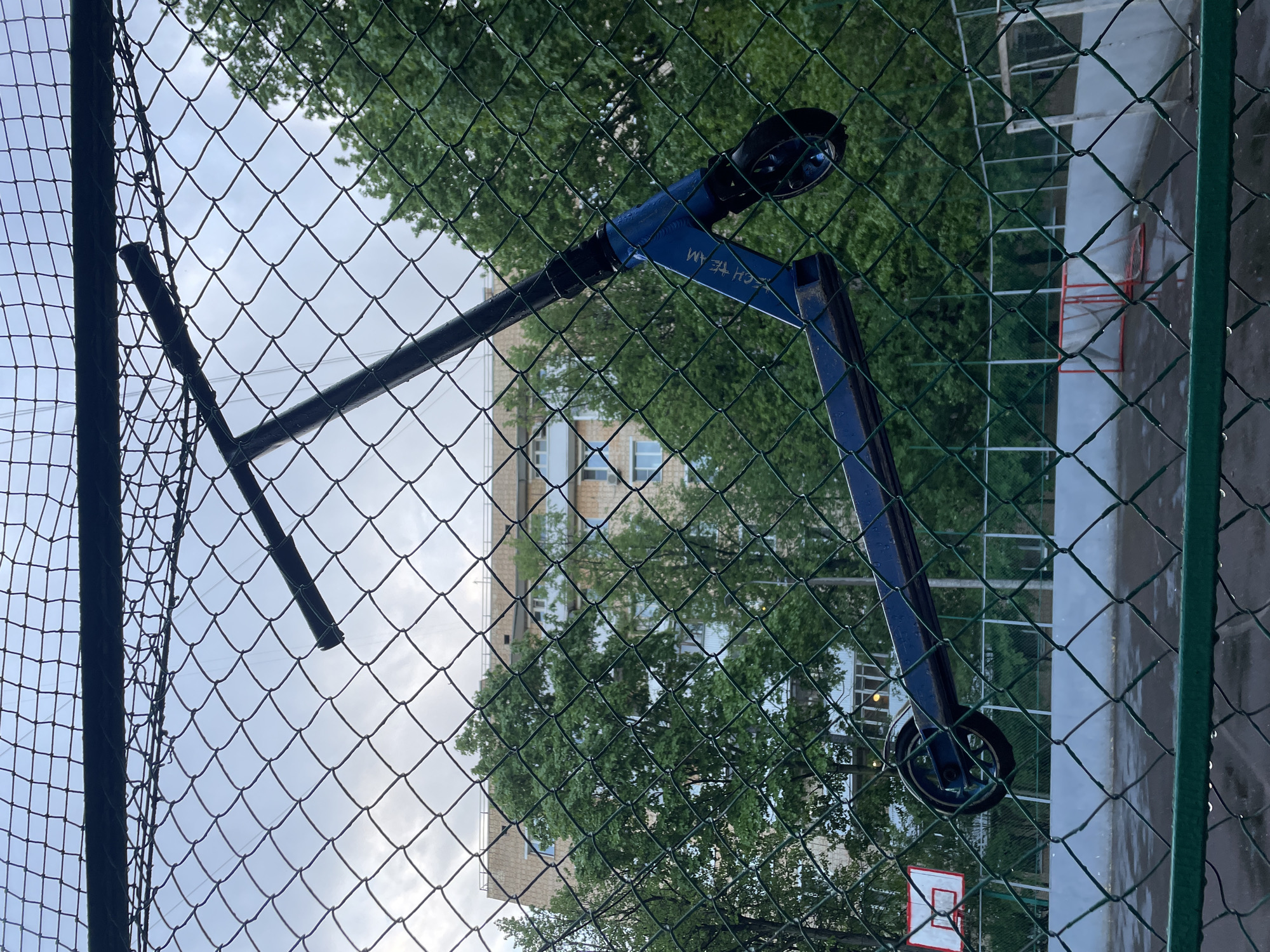 A blue scooter is leaning against a green chain-link fence. The fence is in front of a basketball court with a red-rimmed hoop and wet pavement. Trees and a light-colored building with windows are visible through the fence. The image was taken in Khimki, Russia.