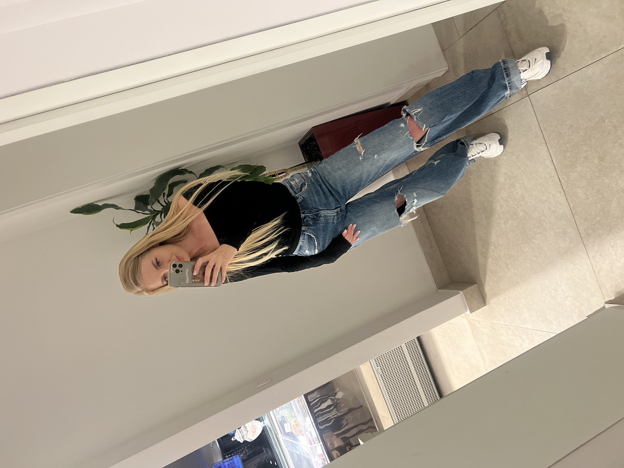 A young woman with long blonde hair is reflected in a mirror. She is wearing a black long-sleeved shirt and ripped light-wash denim jeans. She is holding a cell phone in her right hand, taking a picture of herself. The reflection shows her standing on a light-colored tiled floor. A potted plant is beside her, resting on top of what appears to be a low cabinet or shelf. The overall setting has a modern interior aesthetic, with parts of what appear to be retail displays visible in the reflection.
The image does not provide information to definitively place the photo in Boreti, Montenegro. Therefore, no statement regarding this city can be made.
