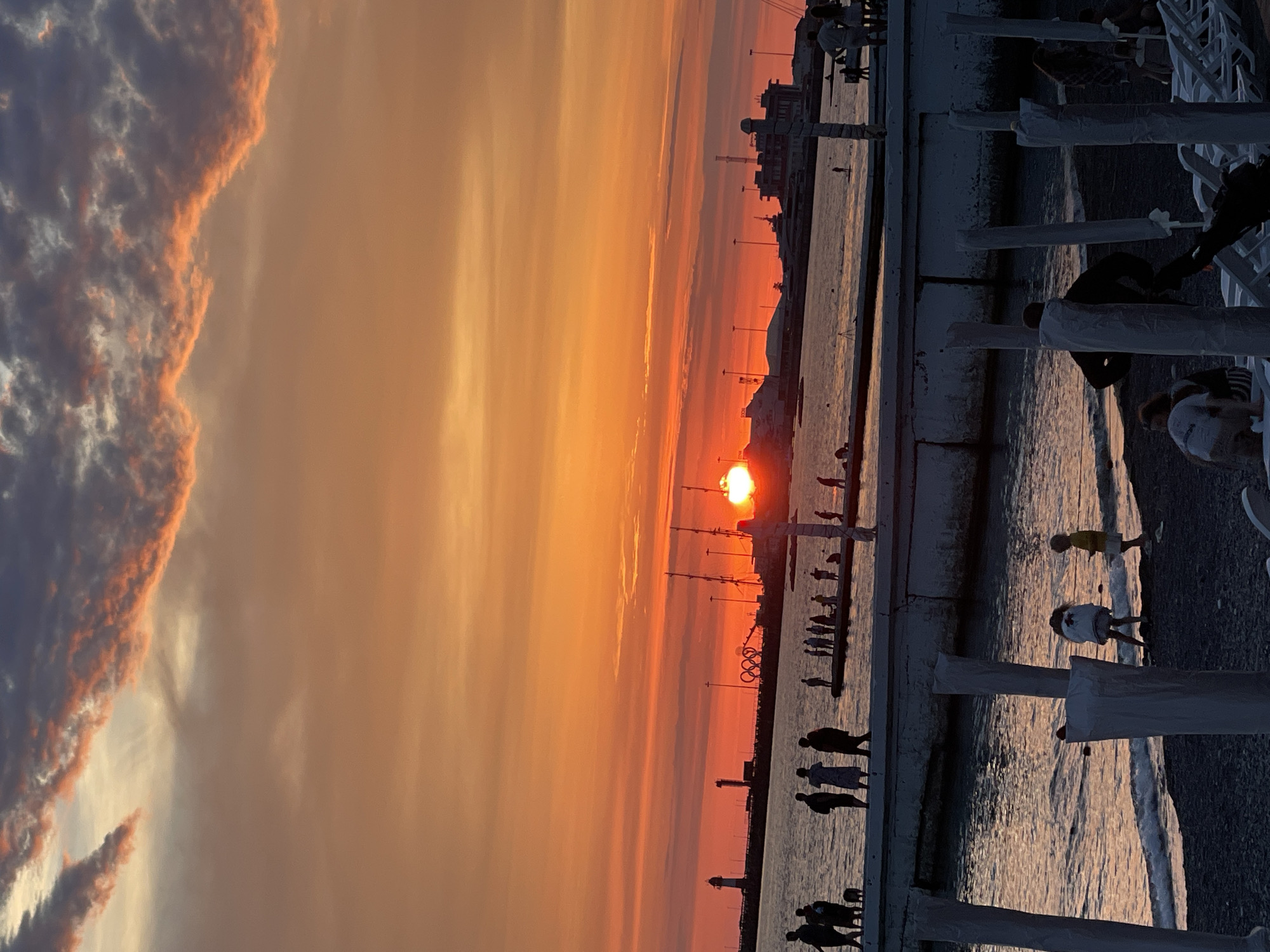 This image depicts a sunset scene at the Sochi Seaport in Sochi, Russia. The sun, a bright orange orb, is setting over the horizon, casting a warm glow across the sky. The sky is filled with a gradient of orange, pink, and purple hues, with some darker clouds adding contrast.
In the foreground, a group of people are walking along a pier, silhouetted against the vibrant sky. Some are strolling casually, while others appear to be engaged in conversation. Further along the pier, more people are gathered, some standing and looking out at the sea, while others are walking.
The sea itself is calm, reflecting the colors of the sunset. In the distance, the Sochi Seaport infrastructure is visible, including lighthouses and several buildings. The iconic Olympic rings are also present, standing tall against the dusky sky, indicating a location related to the 2014 Winter Olympics.
On the beach in the foreground, several figures are visible, including children playing near the water's edge. Beach chairs and umbrellas are set up, suggesting a recreational area. The overall scene is one of a tranquil evening at the seaside, with people enjoying the beautiful sunset.