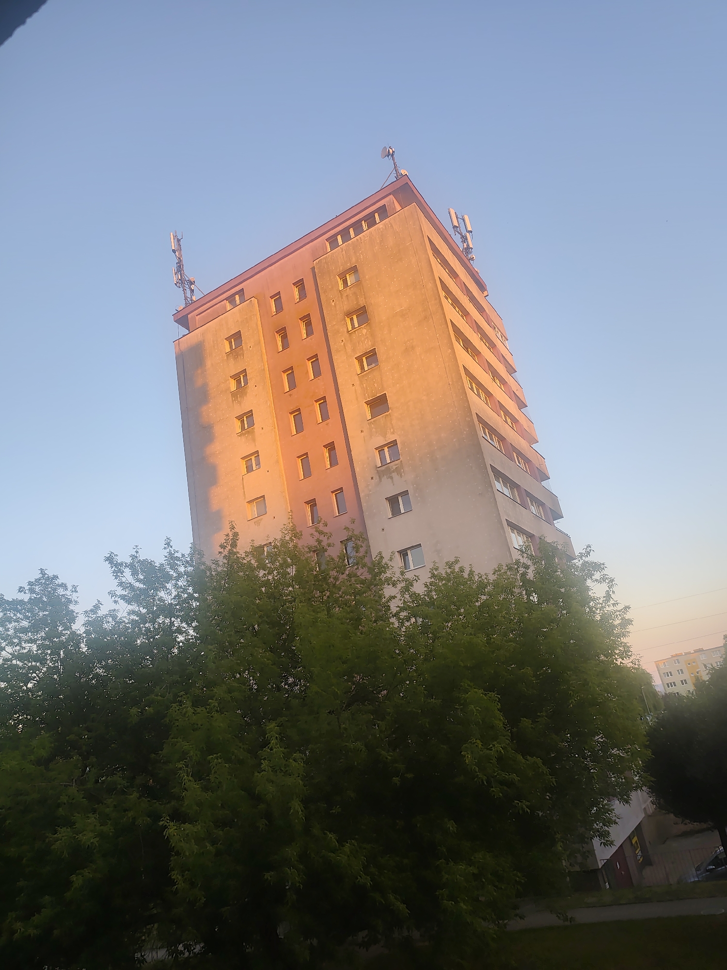 The image depicts a tall, multi-story residential building in Szczecin, Poland, bathed in the warm glow of what appears to be either sunrise or sunset. The building, with its uniform rows of windows, is angled diagonally in the frame. On the roof, several communication antennas are visible, suggesting it might house telecommunication equipment. In the foreground, lush green trees partially obscure the lower floors of the building. The sky is a clear, pale blue, transitioning to a warmer hue near the horizon. In the background, other buildings and parts of the urban landscape are visible, though less detailed. The scene has a peaceful, urban ambiance, characteristic of a residential area in the city.