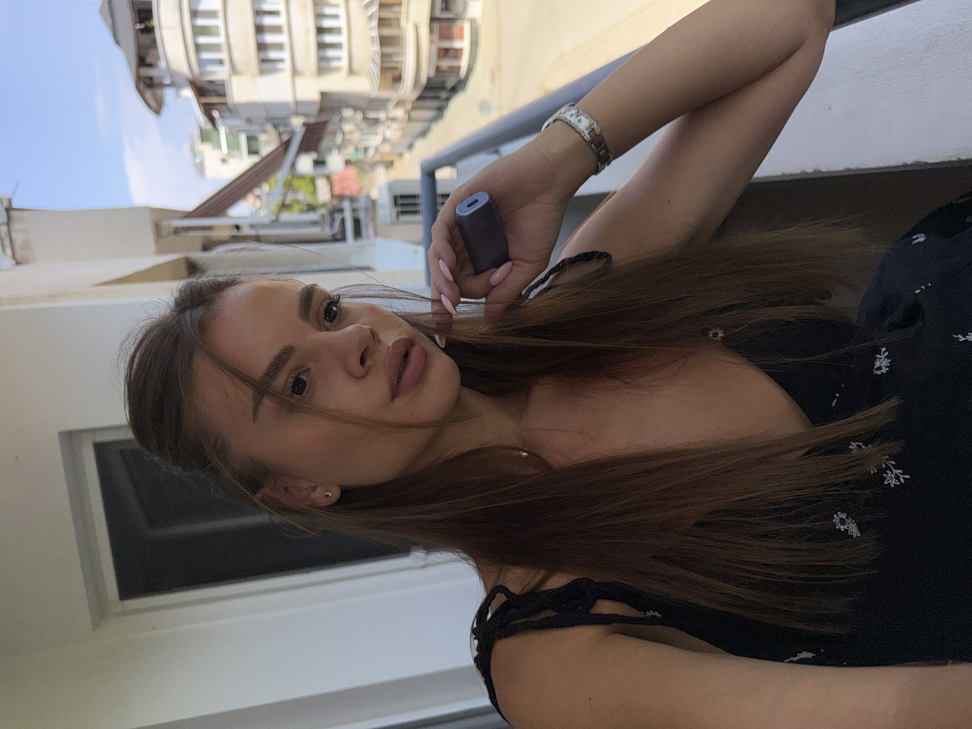 A young woman with long brown hair is standing on a balcony overlooking a street in Plovdiv, Bulgaria. She is wearing a black dress with a floral pattern and holding a dark-colored electronic cigarette device in her right hand. A silver bracelet with white accents is visible on her left wrist. The background shows buildings with balconies and windows, characteristic of urban architecture in Plovdiv. The sky is clear and blue.