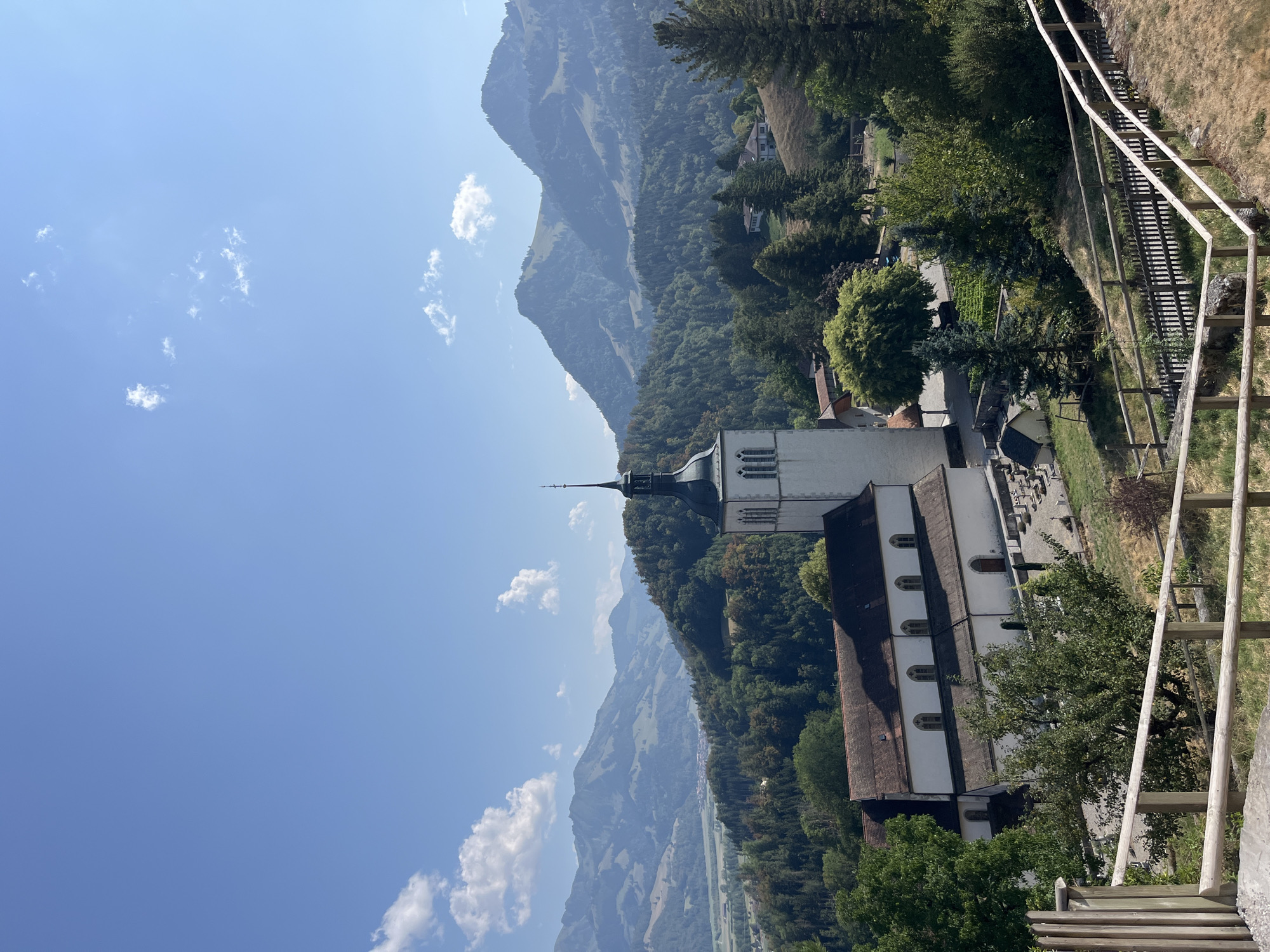The image depicts a landscape scene in the city of Epagny, Switzerland, dominated by a white church with a tall, dark steeple. The church features arched windows and a red-tiled roof. In front of the church, there is a small cemetery with gravestones. Surrounding the church and cemetery are lush green trees and rolling hills. In the background, majestic mountains rise against a clear blue sky with scattered white clouds. The foreground shows a wooden fence and a grassy slope. The overall scene conveys a sense of tranquility and natural beauty.